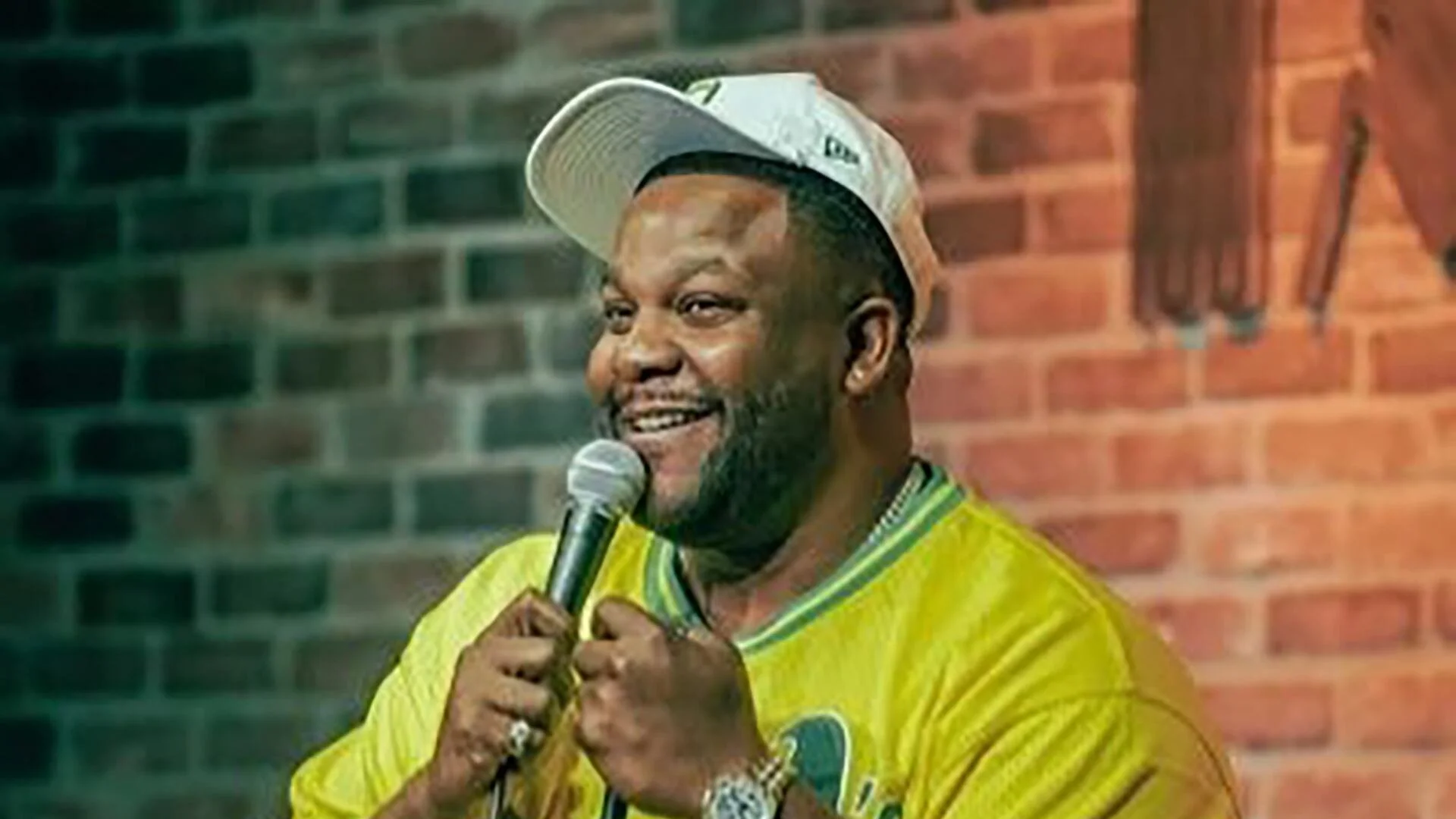 A man wearing a white cap and a bright yellow shirt, smiling while holding a microphone, standing in front of a brick wall.