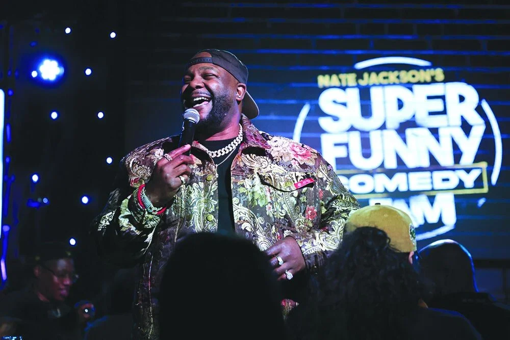 A male stand-up comedian holding a microphone on stage, smiling, wearing a patterned shirt and baseball cap backwards, with a neon sign in the background that says "Matte Jackson's Super Funny Comedy Show". Audience members are visible in the foreground.