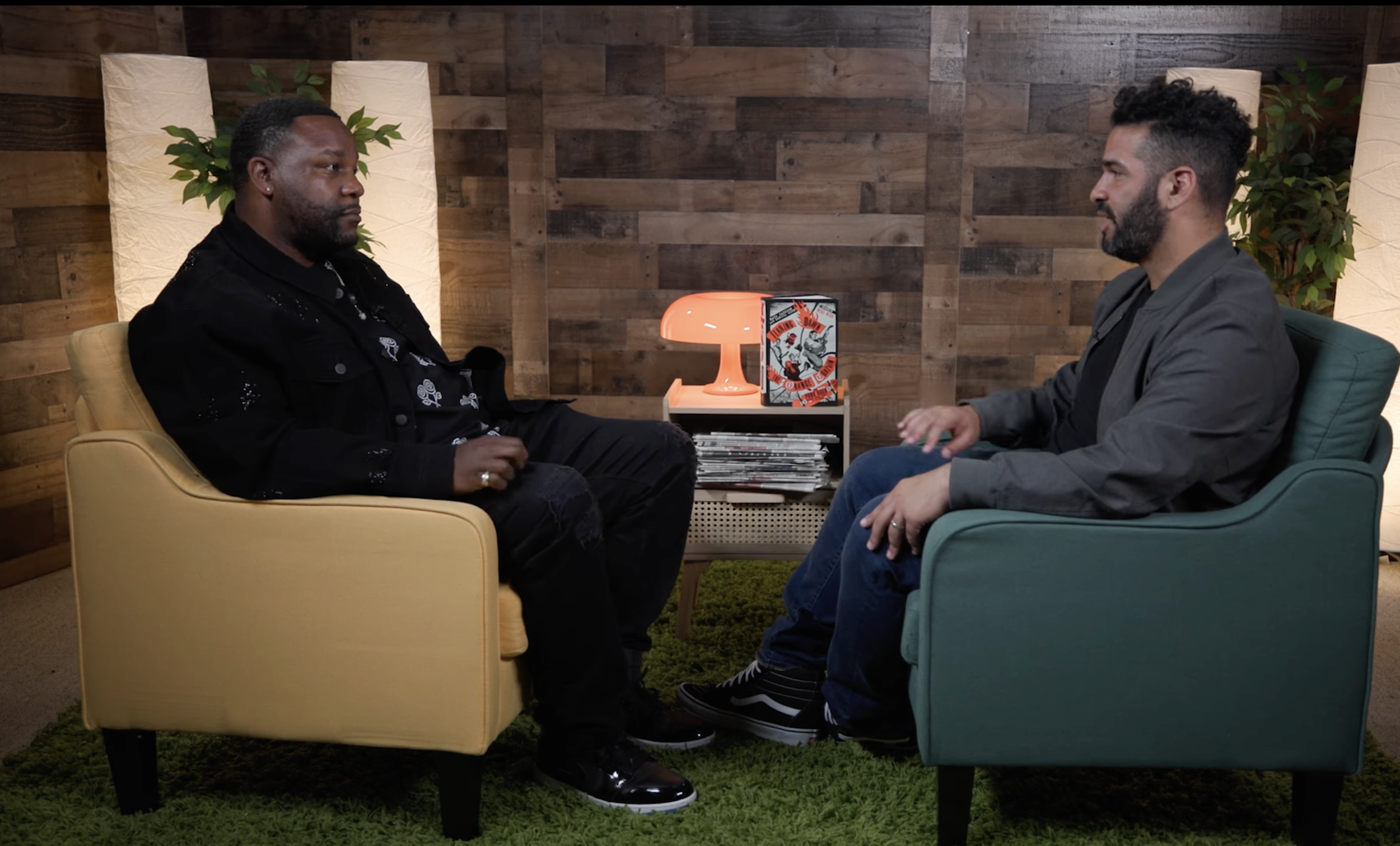 Two men are sitting across from each other in a cozy room with wood-paneled walls, engaged in conversation. One man is wearing a black jacket and black pants, while the other man is wearing a gray blazer and blue jeans. There is an orange lamp and a collection of magazines on a small table between them, with some potted plants in the background.