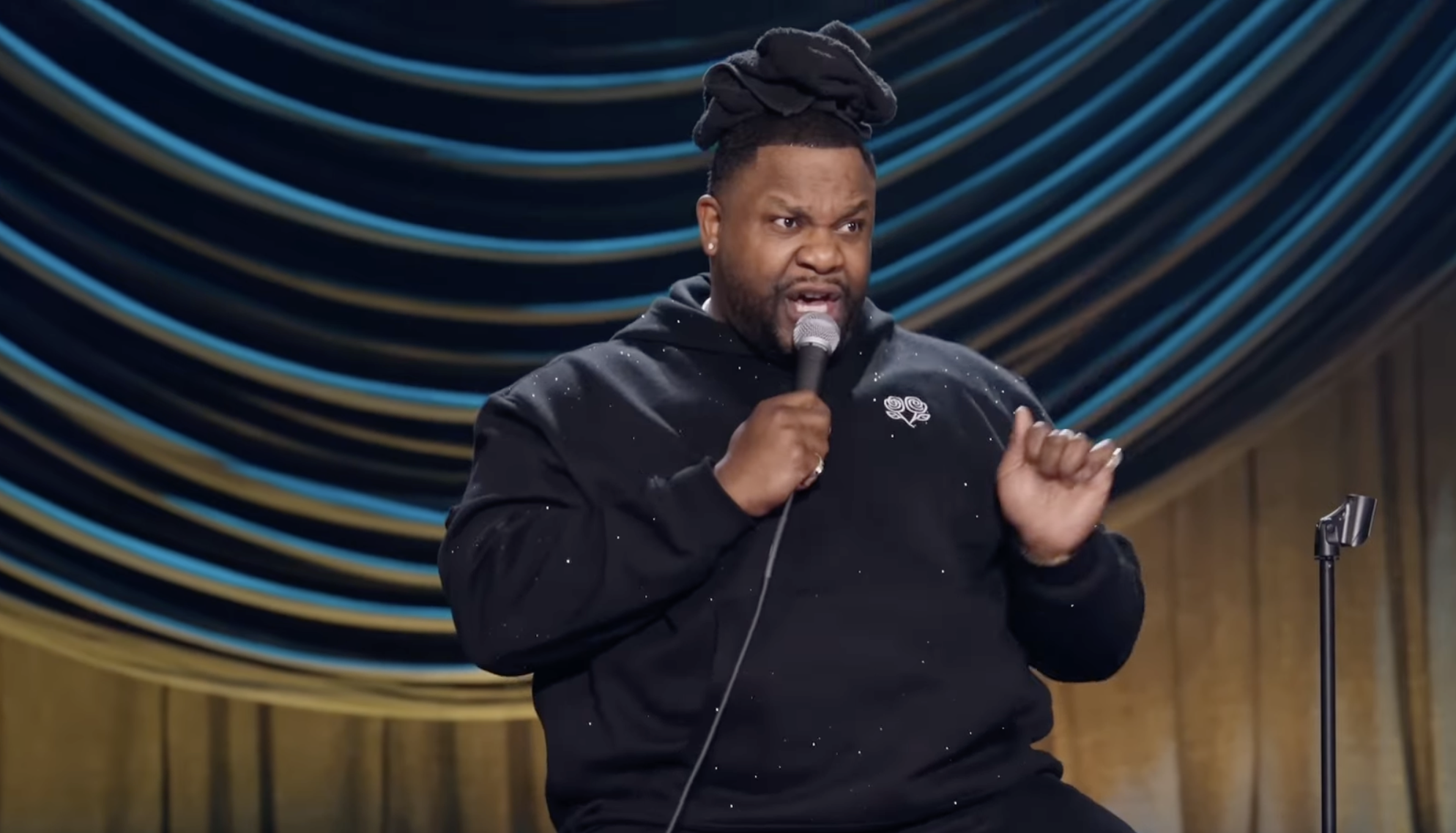 A man with dreadlocks styled in a bun, wearing a black hoodie with a small emblem on the chest, speaking into a microphone on a stand, standing in front of a backdrop with blue and tan concentric circular patterns.