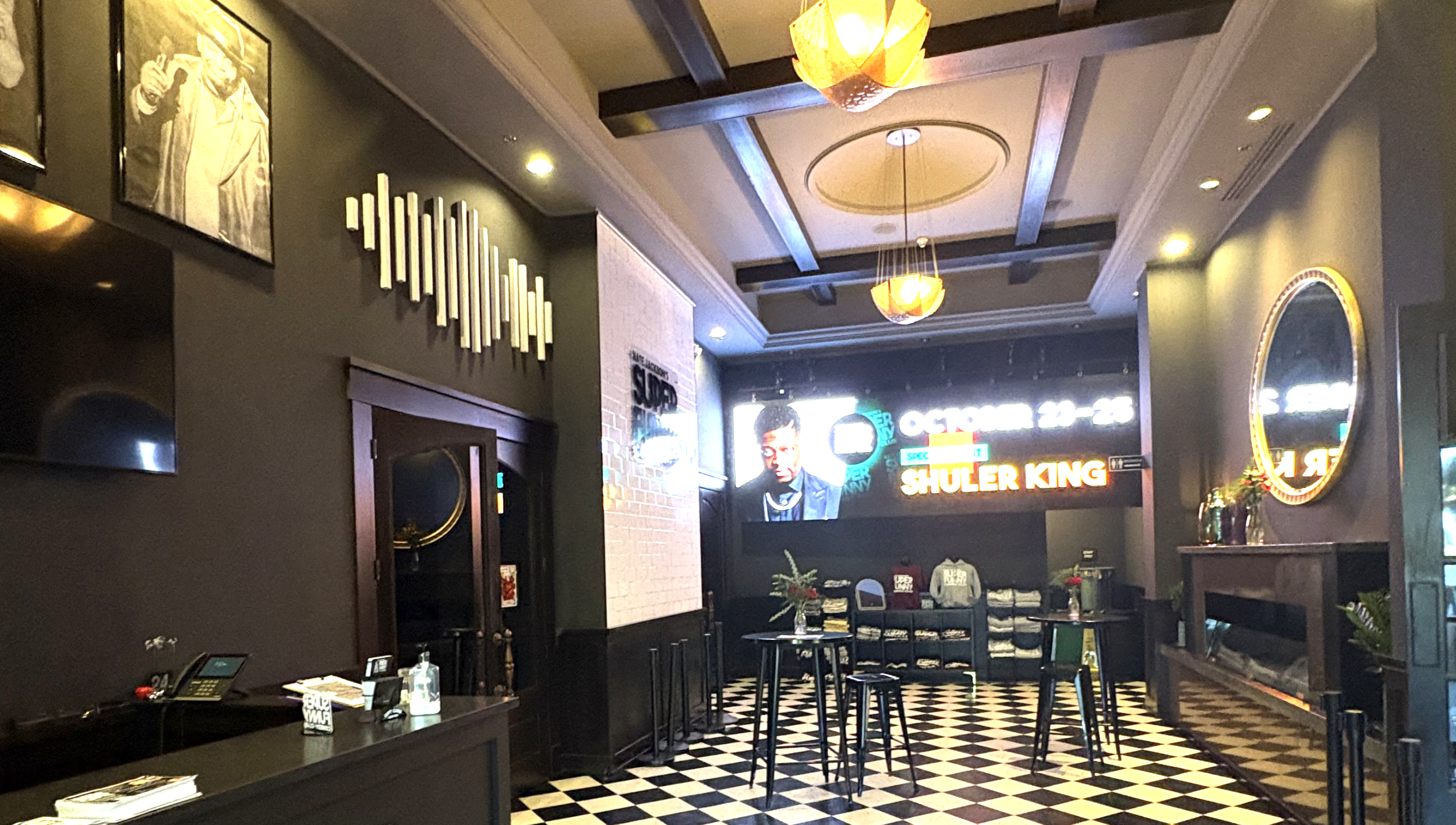 Inside a restaurant or bar with black and white checkered floor, modern lighting fixtures, a large screen displaying an image of Shuler King, a neon sign with the same name, and a few tables and chairs decorated with small flower arrangements.
