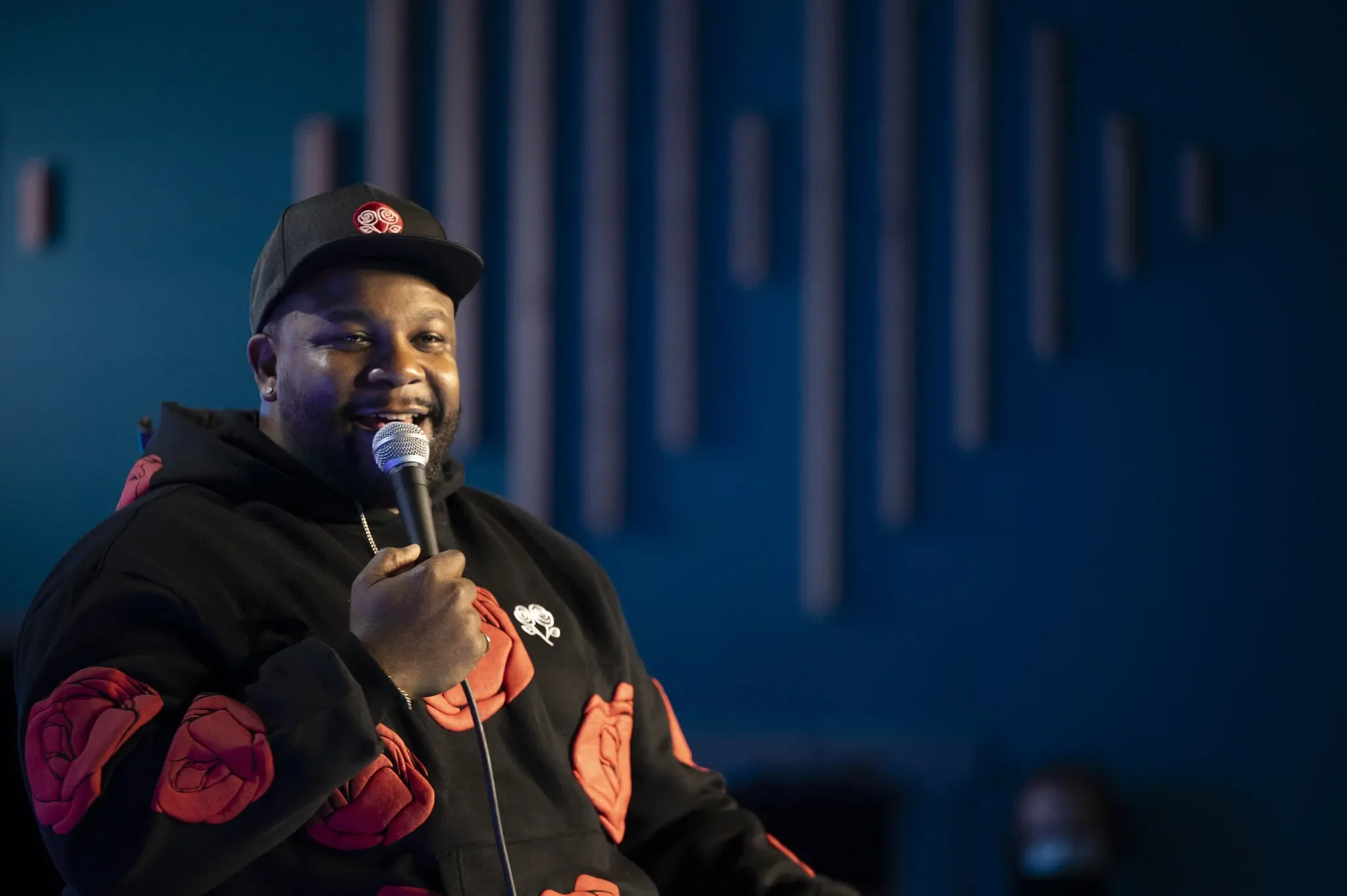 A smiling man holding a microphone, wearing a black cap and a jacket with red details, standing against a blue background with vertical poles.