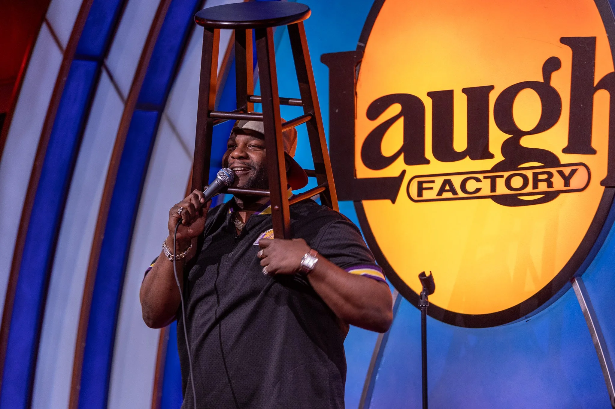 A man holding a microphone on stage, standing behind a chair, in front of a bright orange sign that says 'Laugh Factory'.