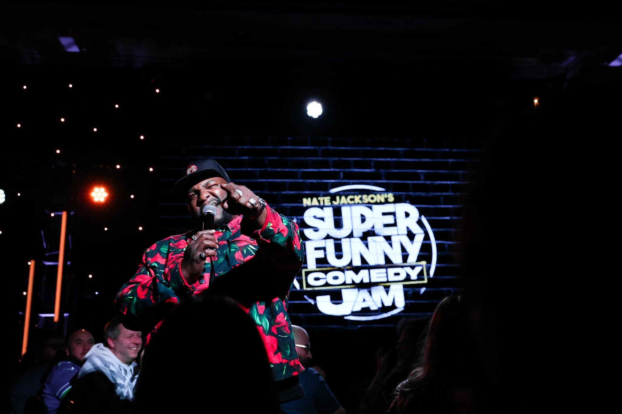A comedian on stage at a comedy show, holding a microphone and pointing towards the camera. Behind him is a large illuminated sign that reads "Nate Jackson's Super Funny Comedy Jam". The audience is visible in the foreground, smiling and enjoying the