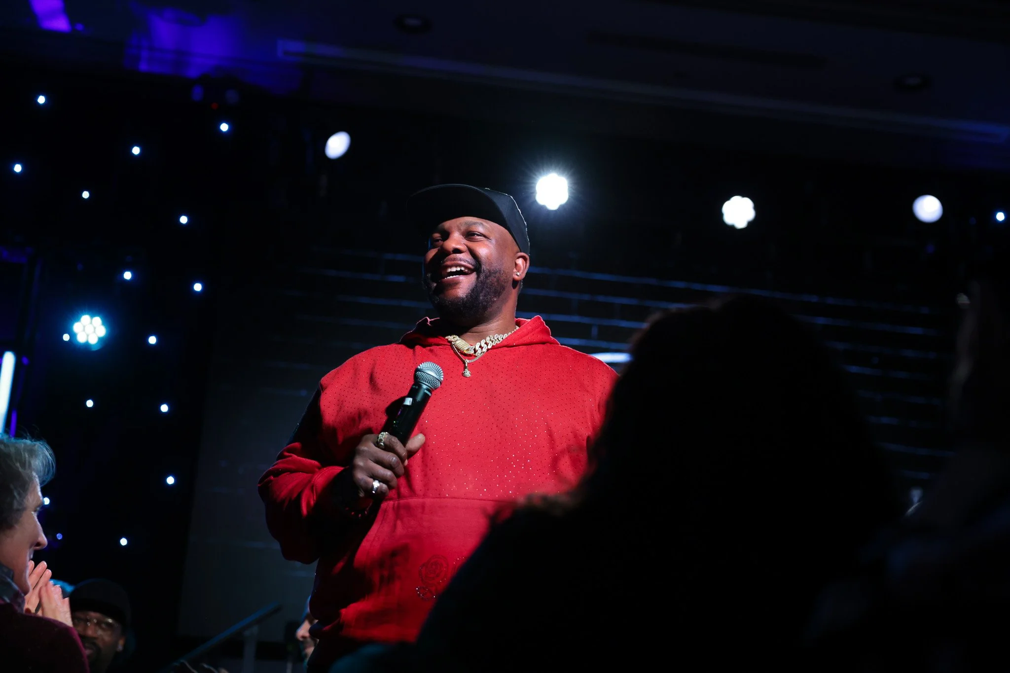 A man wearing a black cap, red hoodie, and gold chains is smiling and holding a microphone on stage with dark background and bright stage lights.