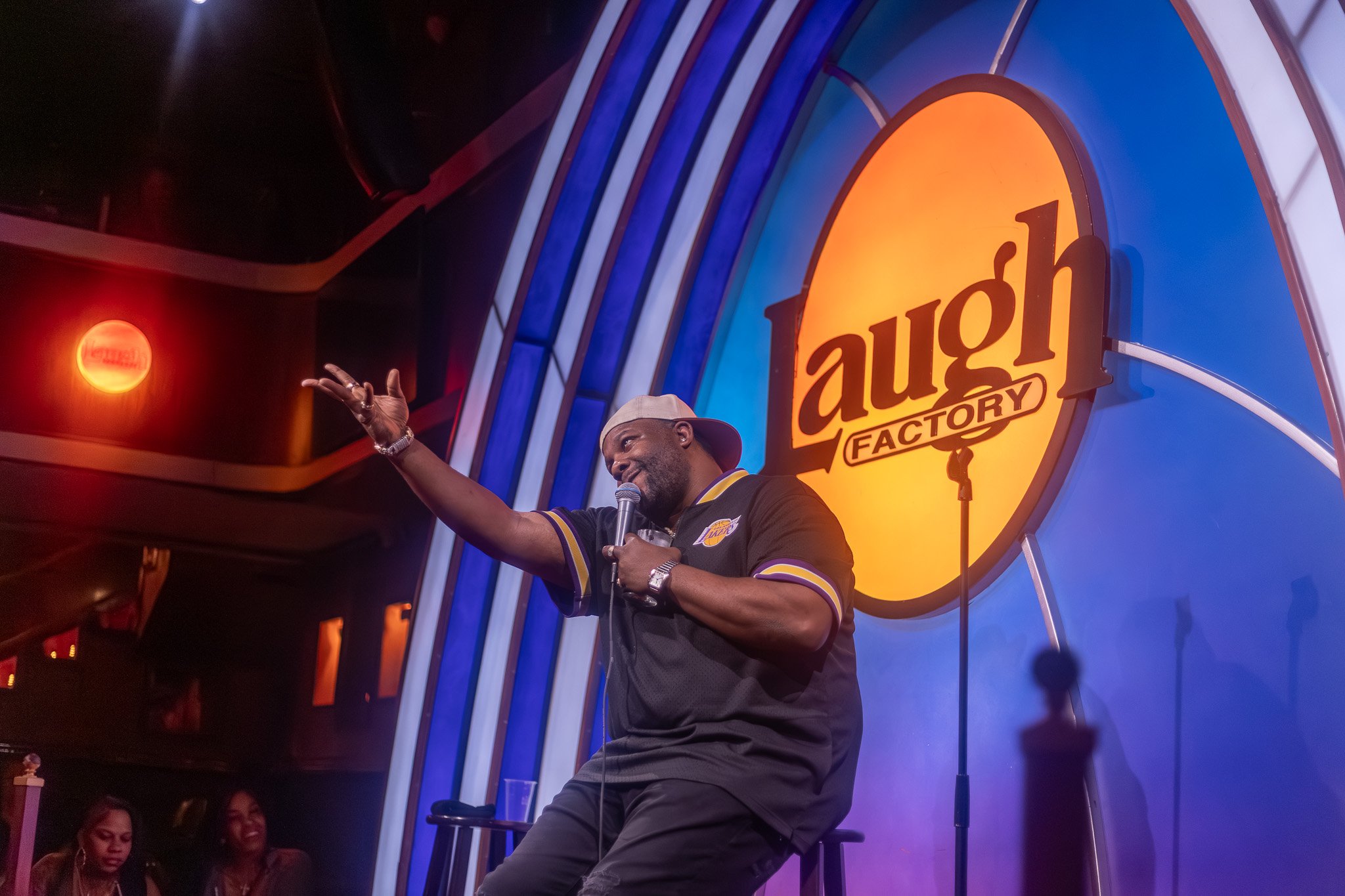 A man wearing a Los Angeles Lakers shirt and a baseball cap, holding a microphone, speaking on stage at a comedy show inside the America's Got Talent studio. The background features the America's Got Talent logo and colorful lights.