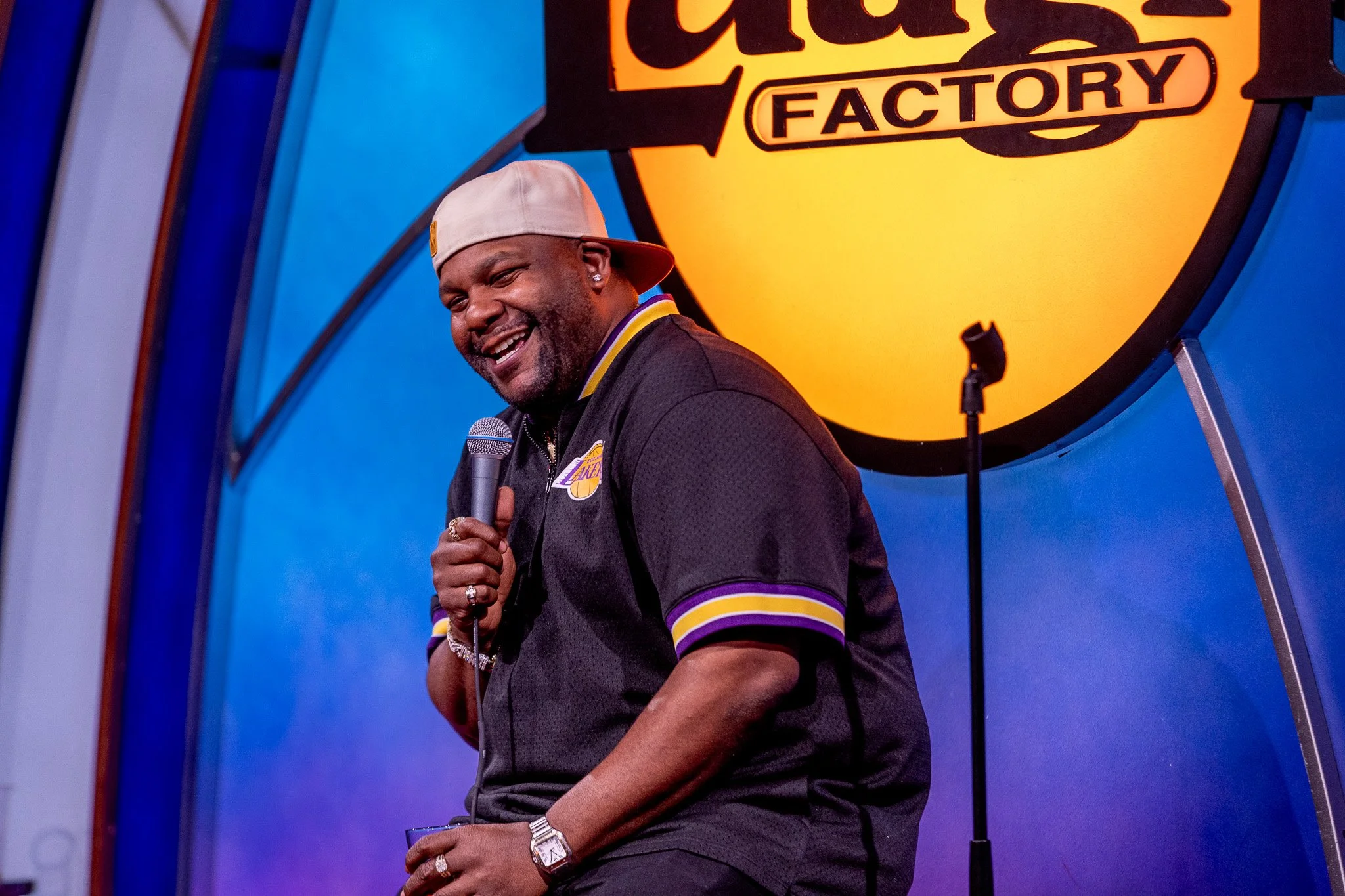 A man smiling while holding a microphone on stage with a yellow logo behind him that reads 'Catch Factory'.