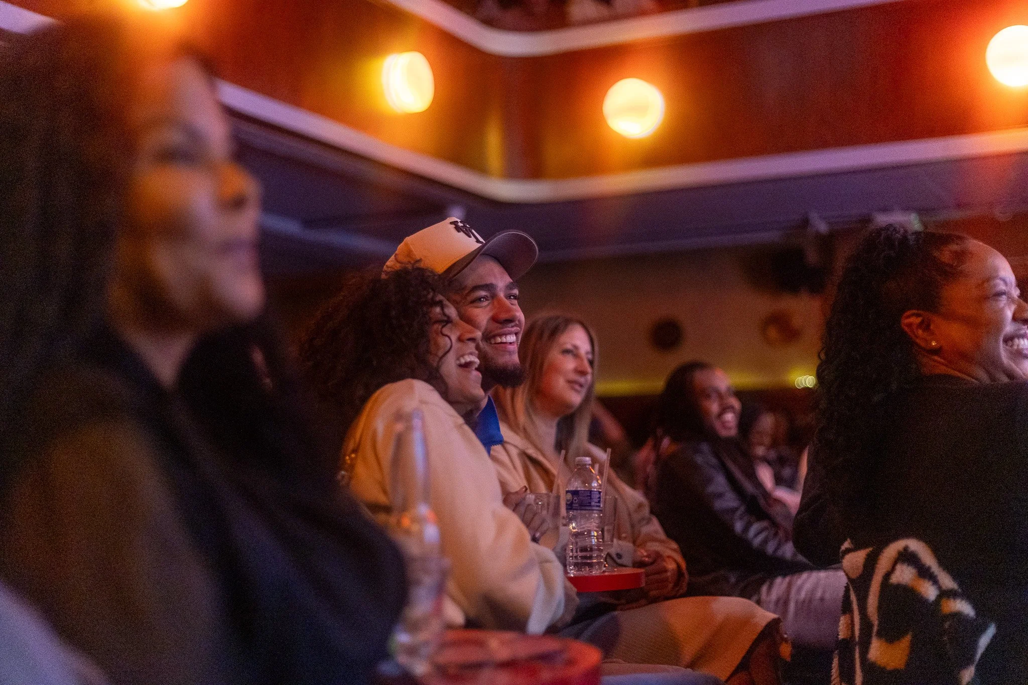Audience of diverse people watching a performance on stage, smiling, and enjoying the show in a dimly lit theater with warm lighting.