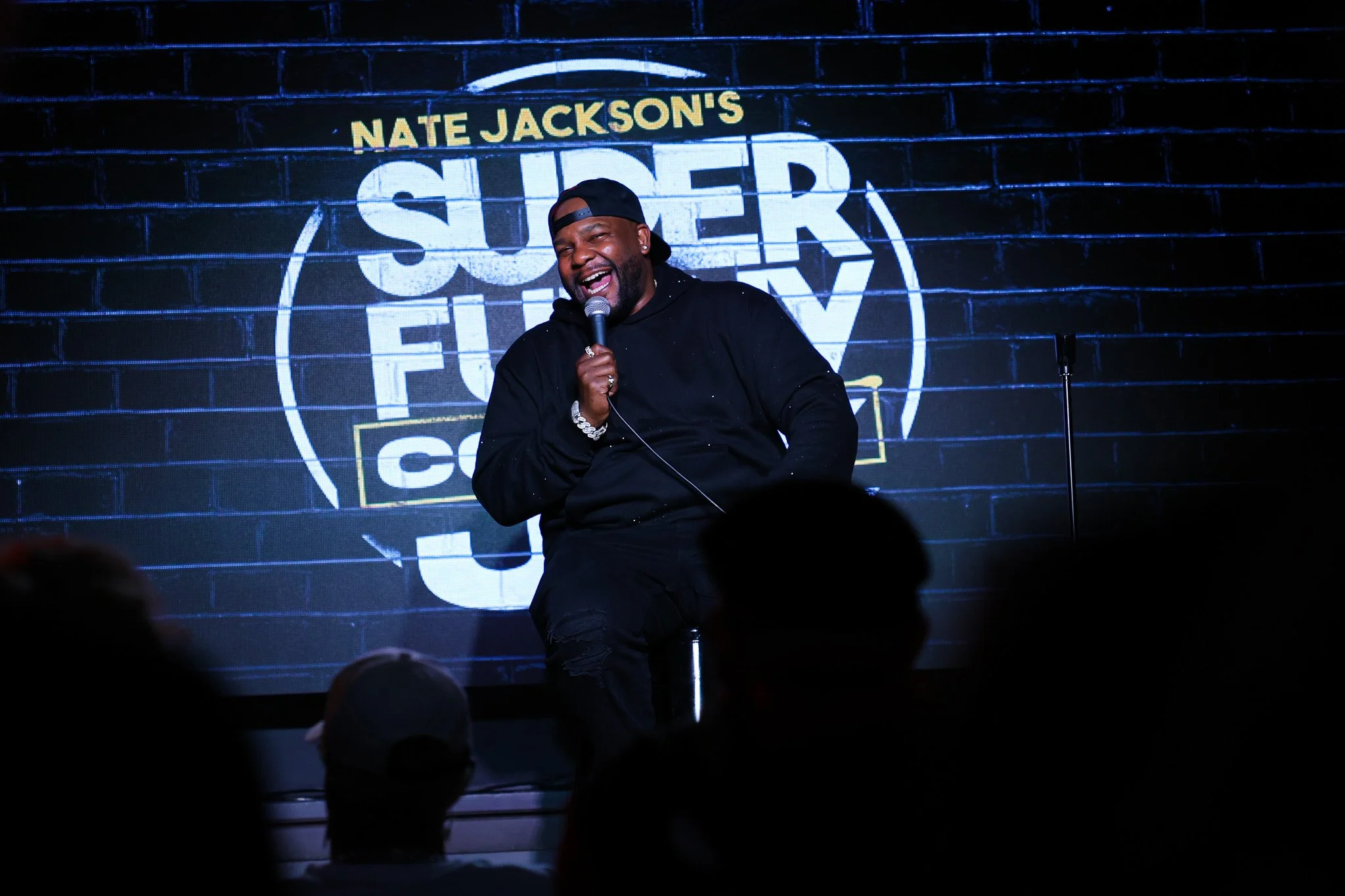 A man sitting on a stage, laughing and holding a microphone, with a sign behind him reading 'Nate Jackson's Super F...' on a brick wall background.