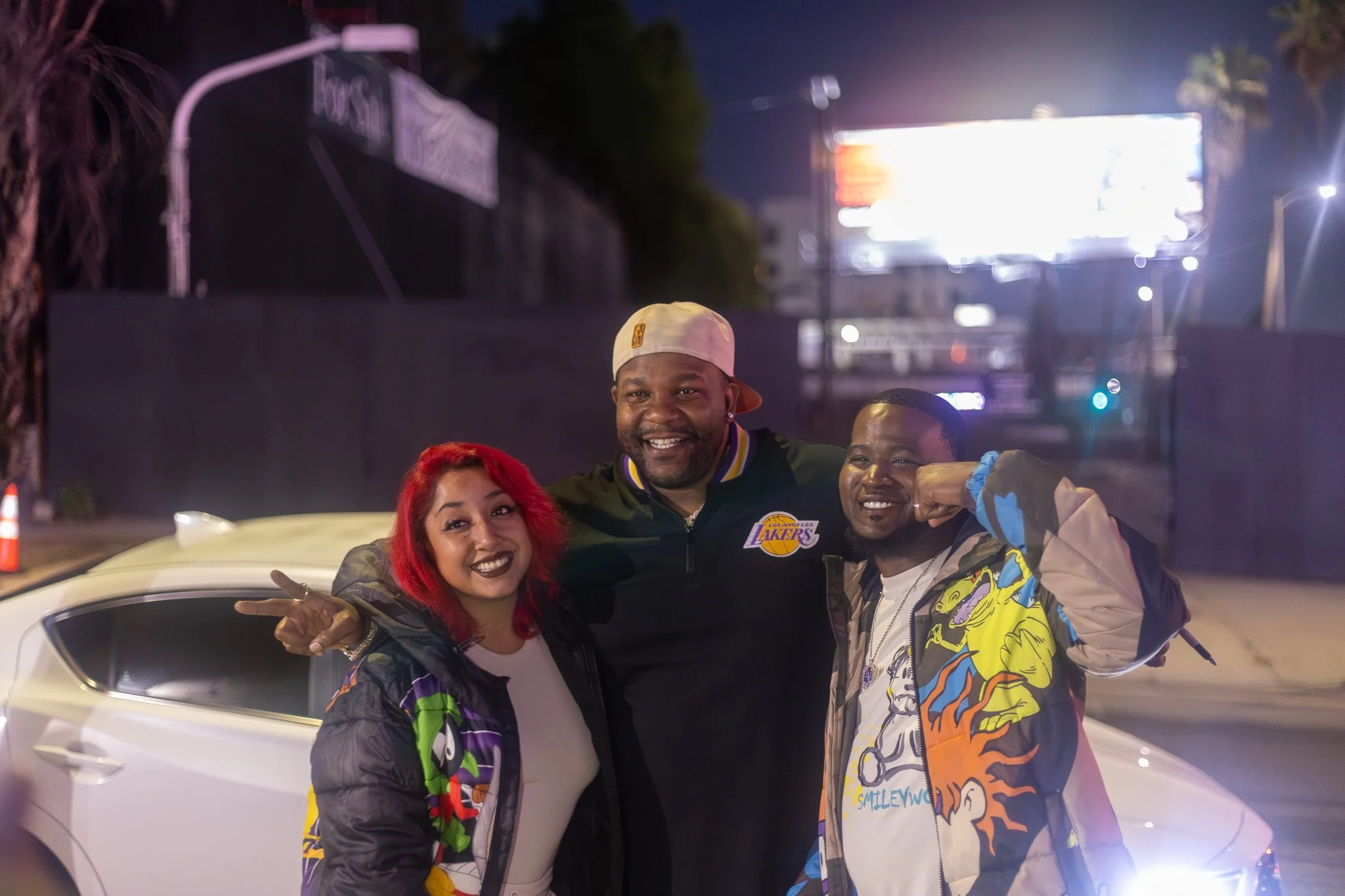 Four people posing together outside at night, standing in front of a white car, with a brightly lit billboard in the background.