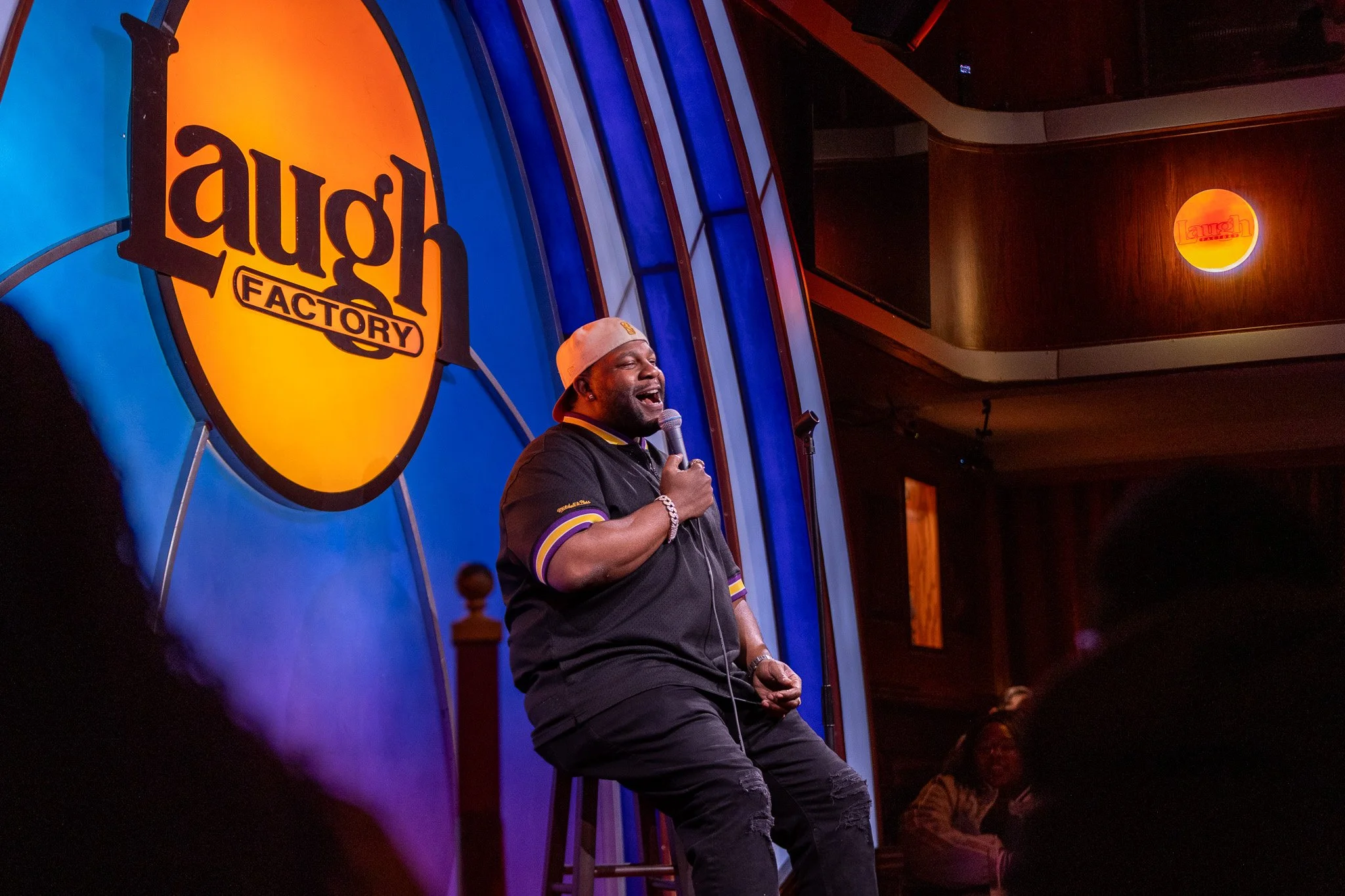 A man holding a microphone on stage at Laugh Factory comedy club, with a bright Laugh Factory sign in the background.