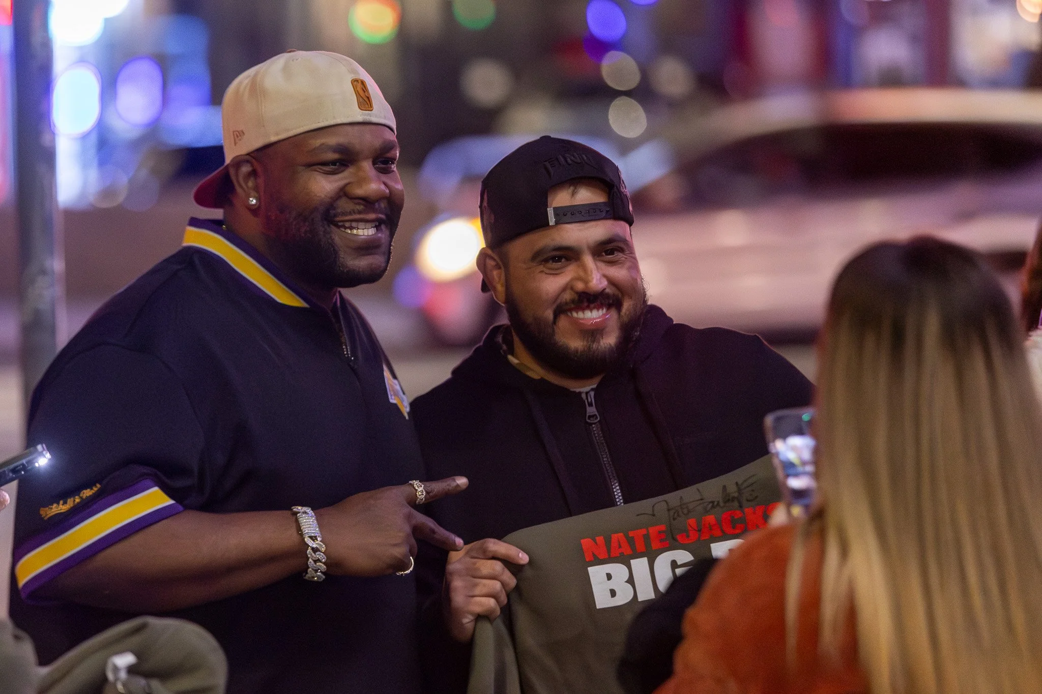 Two men smiling for a photo during a public event at night. One man is wearing a white cap backwards, earrings, a black jacket with purple and yellow trim, and jewelry. The other man is wearing a black cap backwards, a black zip-up hoodie, and holdin