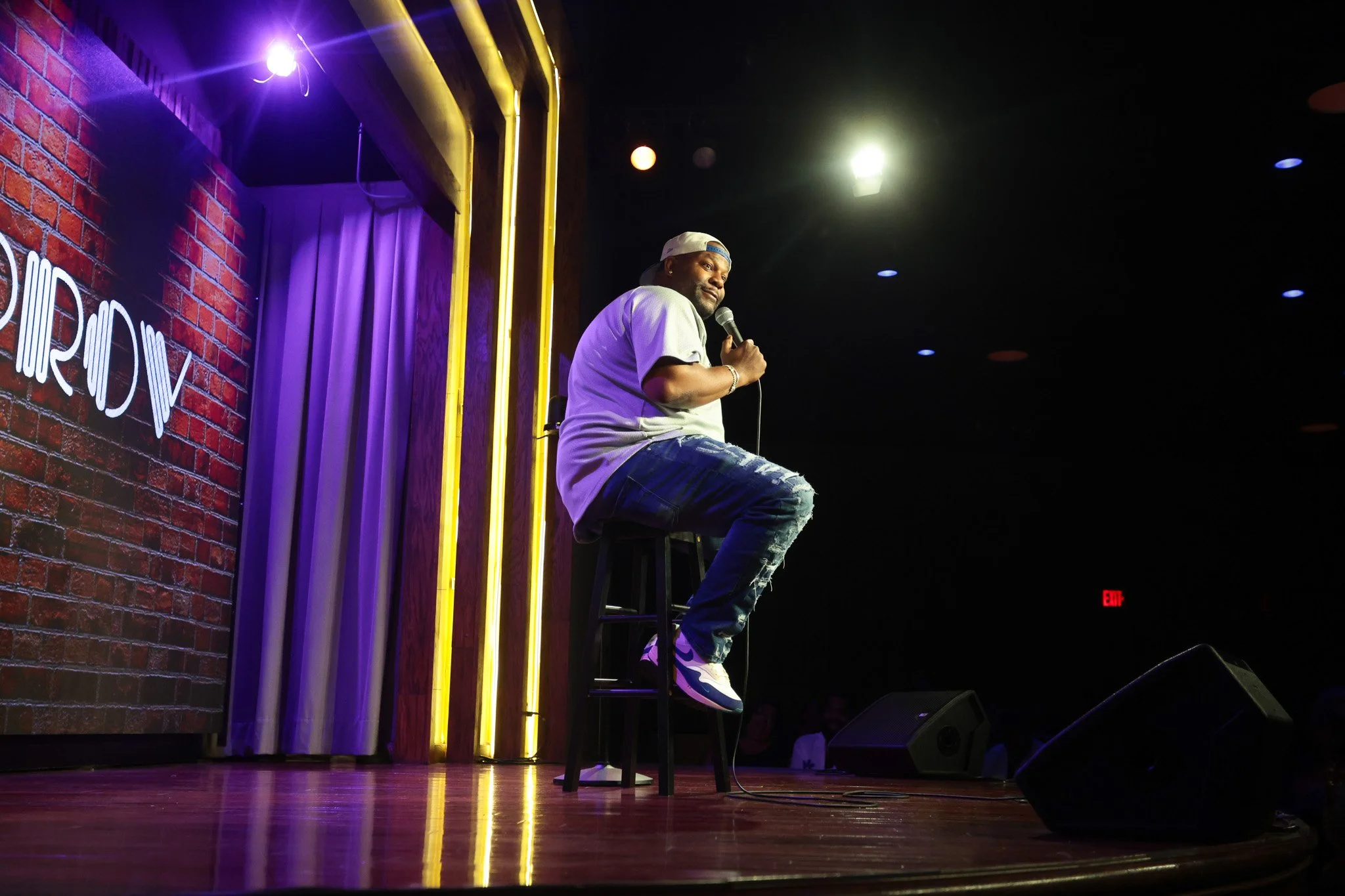 A man performing stand-up comedy on stage with a microphone, sitting on a stool, in a dimly lit comedy club with neon and purple lighting, brick wall, and curtain backdrop.