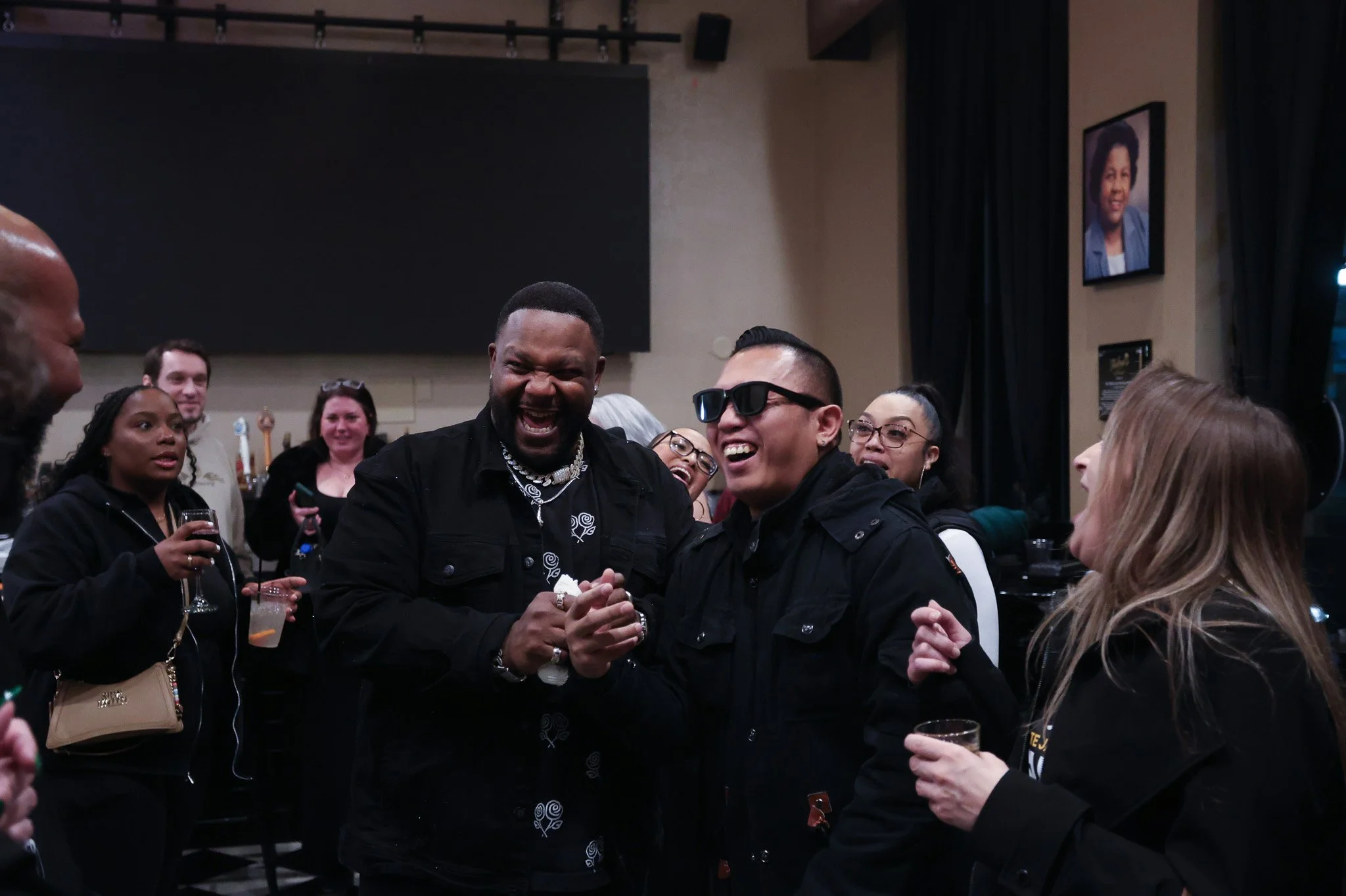 Group of diverse people at a social gathering, smiling and holding drinks, with a man wearing sunglasses at the center.