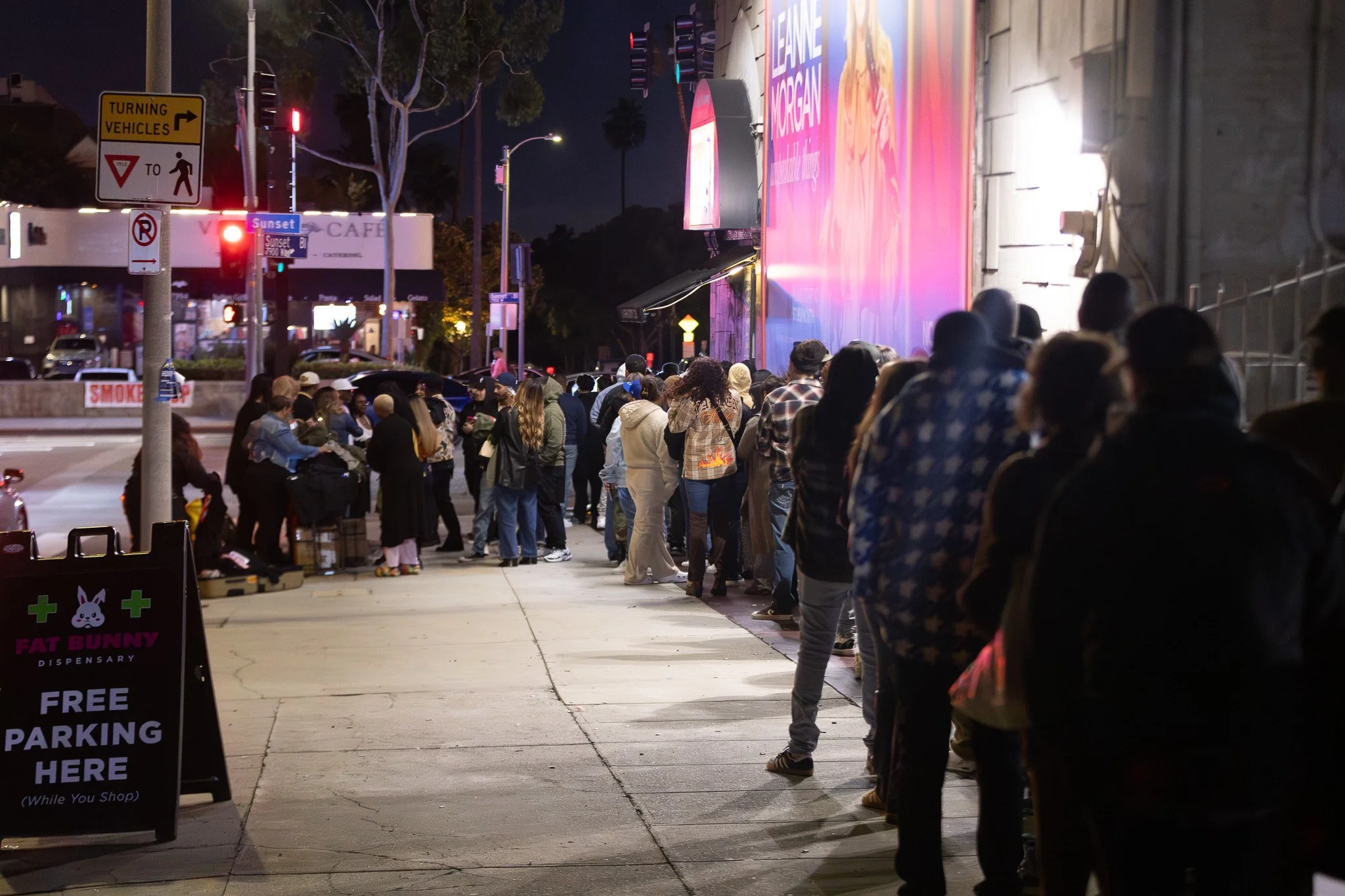 A line of people waiting outside a storefront at night. There are various signs, including one for a dispensary and a sign for free parking. The store has a pink and purple illuminated advertisement.