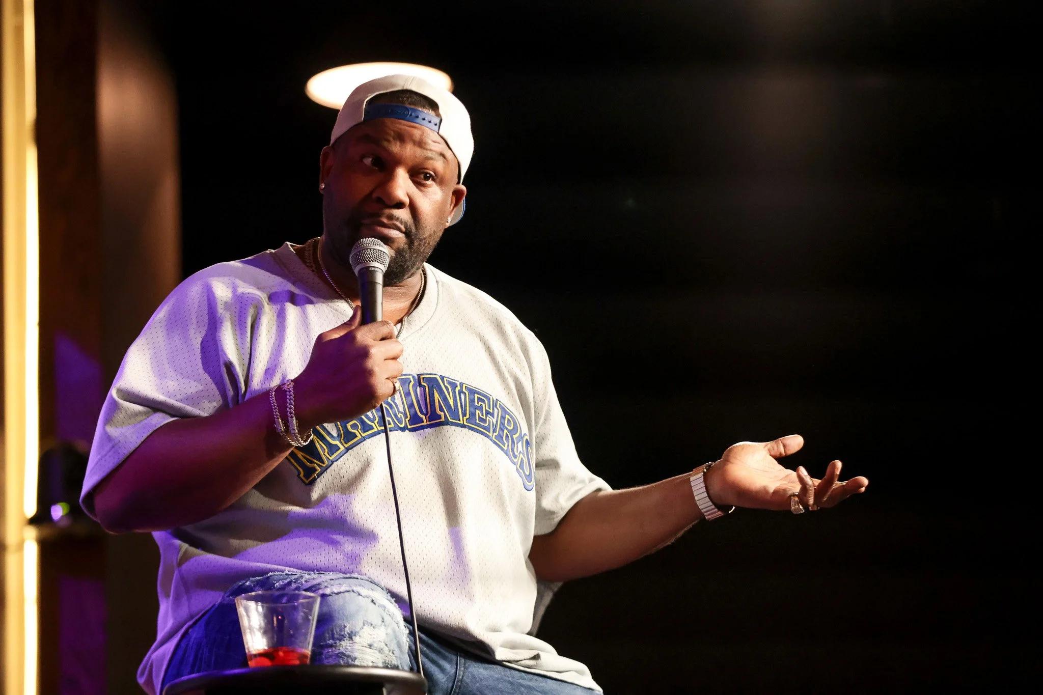A man wearing a white baseball cap backward and a gray hoodie, holding a microphone, speaking on stage with a black background.
