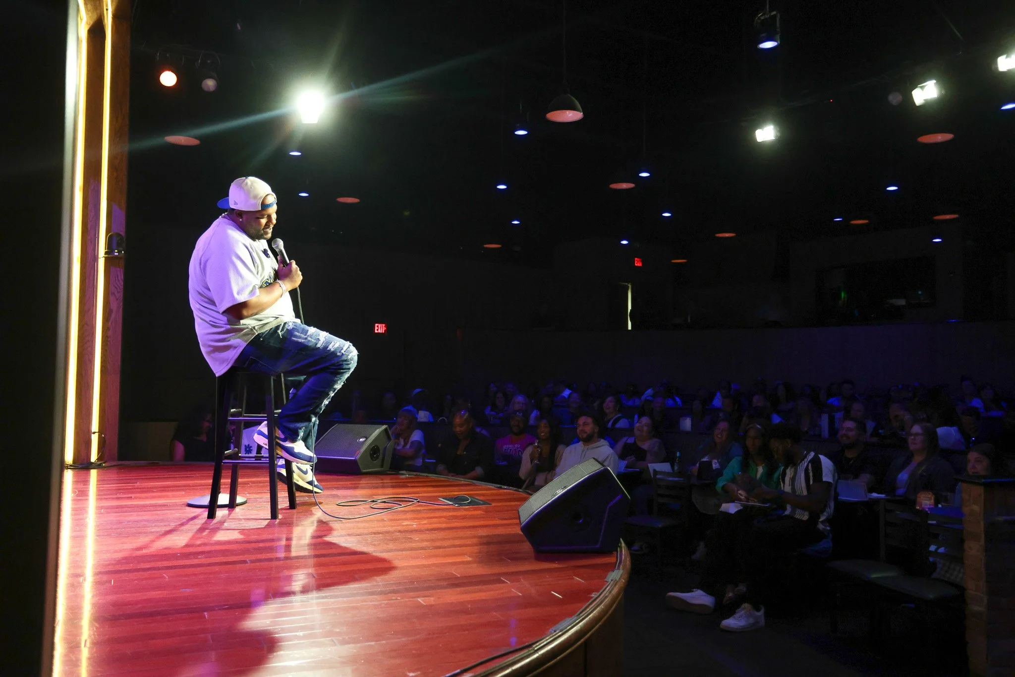 A male comedian performing stand-up comedy on stage at a comedy club. He is sitting on a stool, holding a microphone, wearing a baseball cap backwards, a white t-shirt, and ripped jeans. The stage has bright lighting and the audience is laughing and 