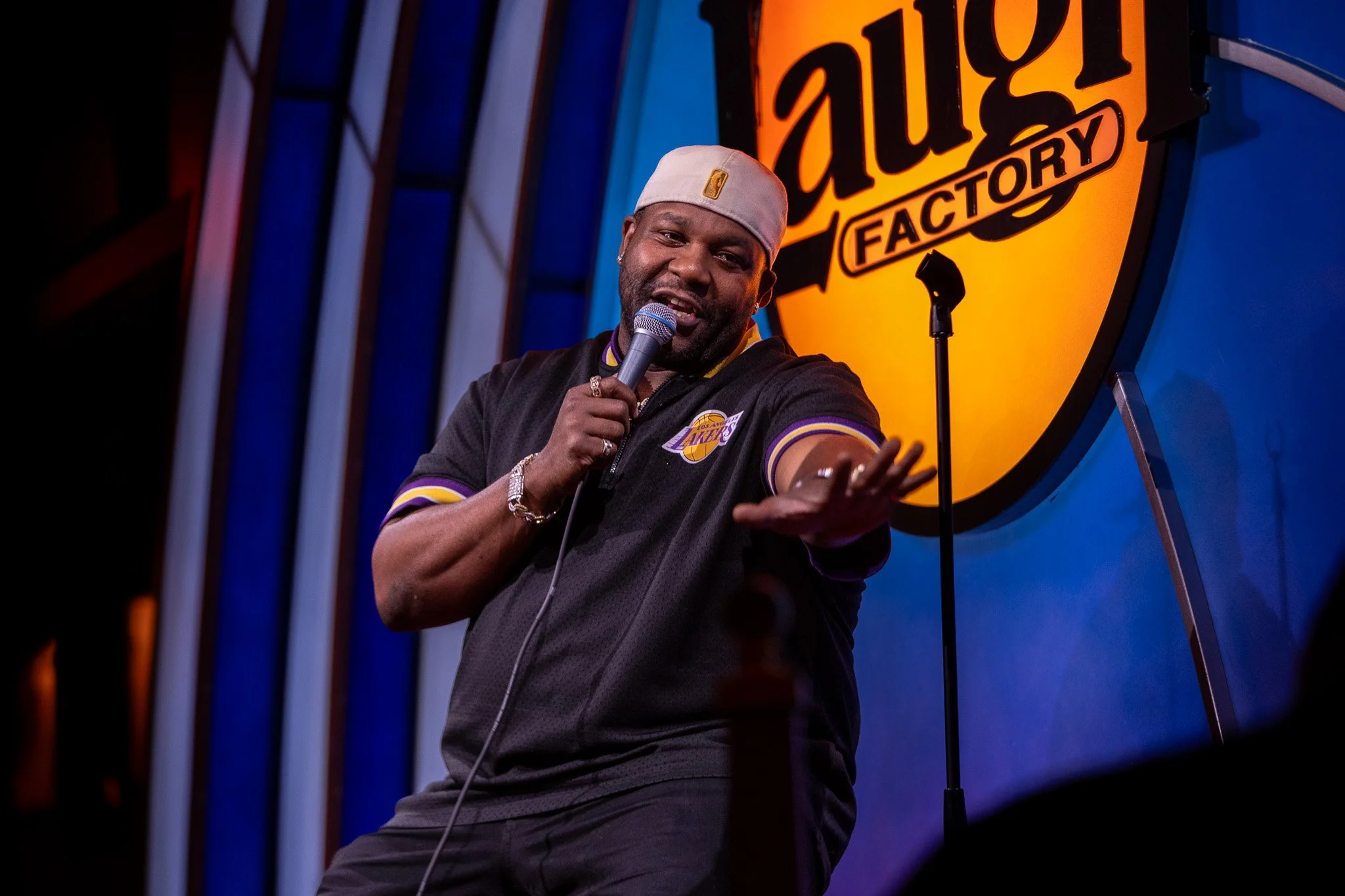 A man on stage at the Laugh Factory comedy club, holding a microphone, wearing a beige hat, a black Los Angeles Lakers jacket, and smiling.
