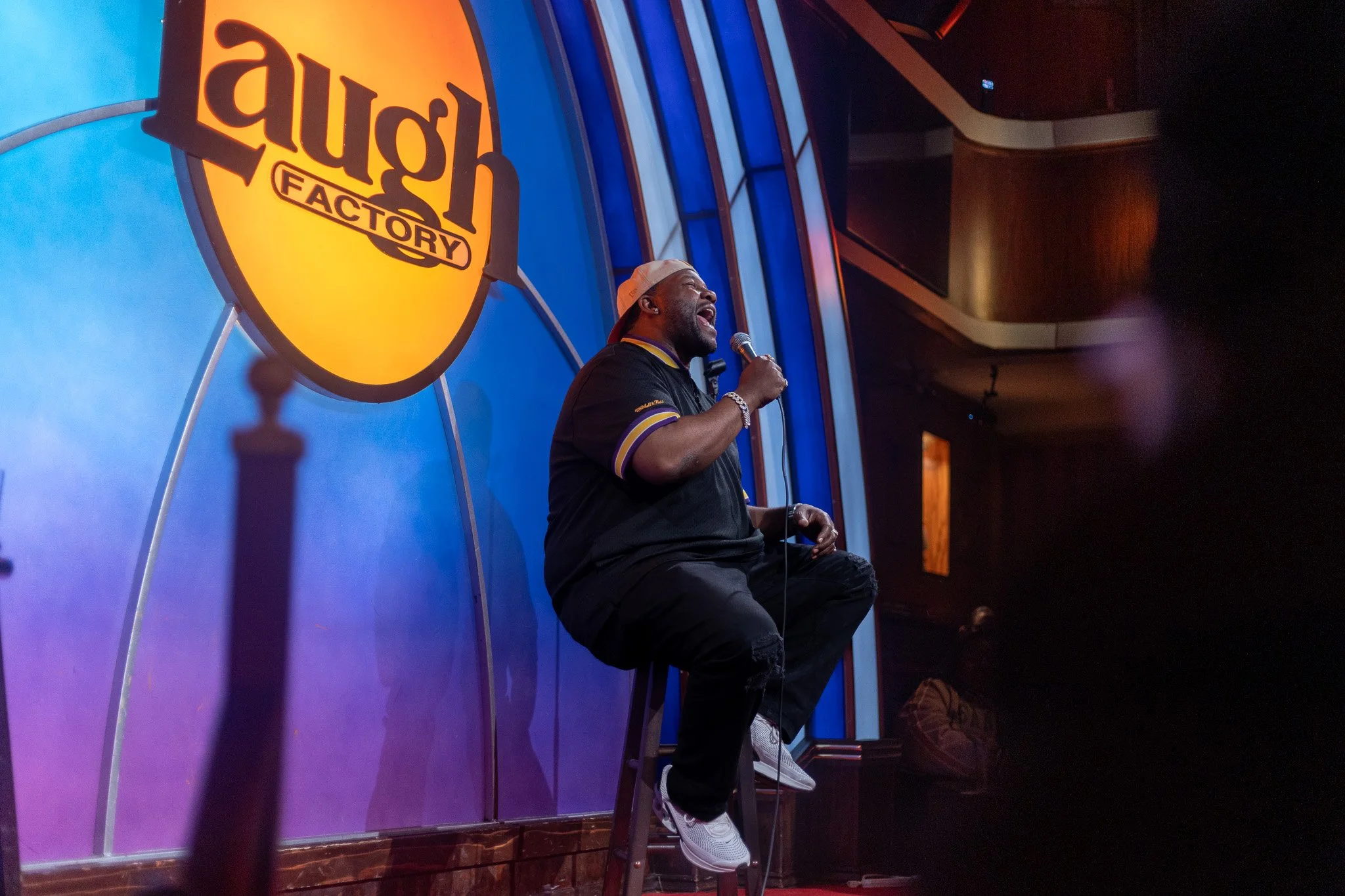 A man sitting on a stool performing stand-up comedy on stage with a microphone in hand, illuminated by stage lights, in front of a large sign that reads 'laugh FACTORY'.
