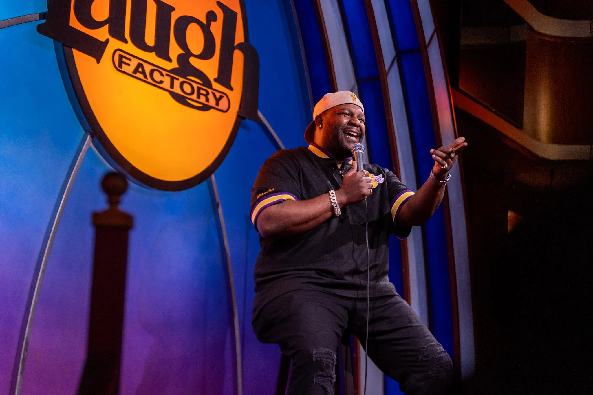 Person in a black and purple sports jersey, wearing a beige baseball cap, holding a microphone and speaking on stage at a comedy show. A large sign with the words 'Laugh Factory' is visible in the background.