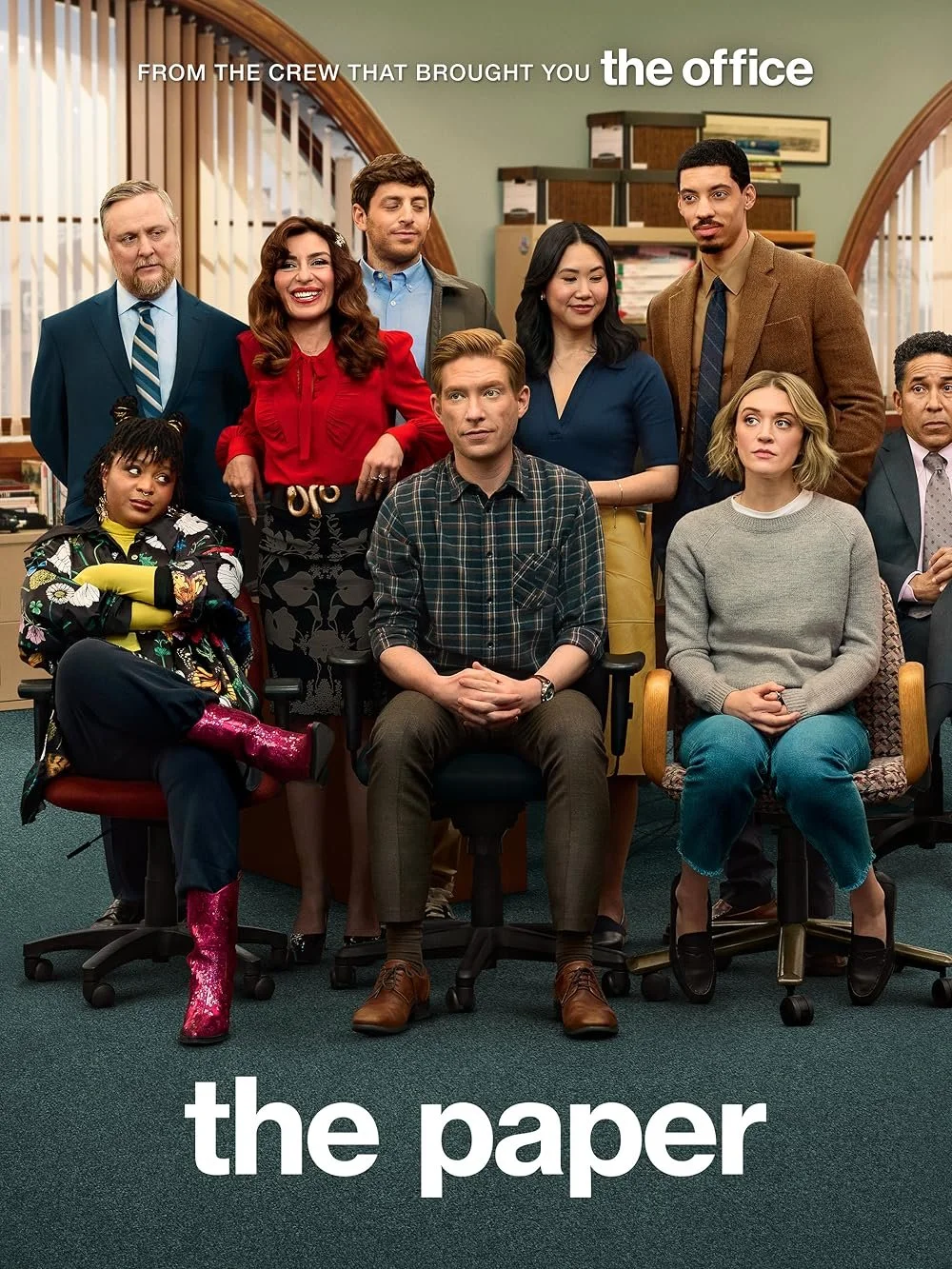 Group photo of diverse office workers in a conference room with green walls and bookshelves, with the text "from the crew that brought you the office" at the top and "the paper" at the bottom.