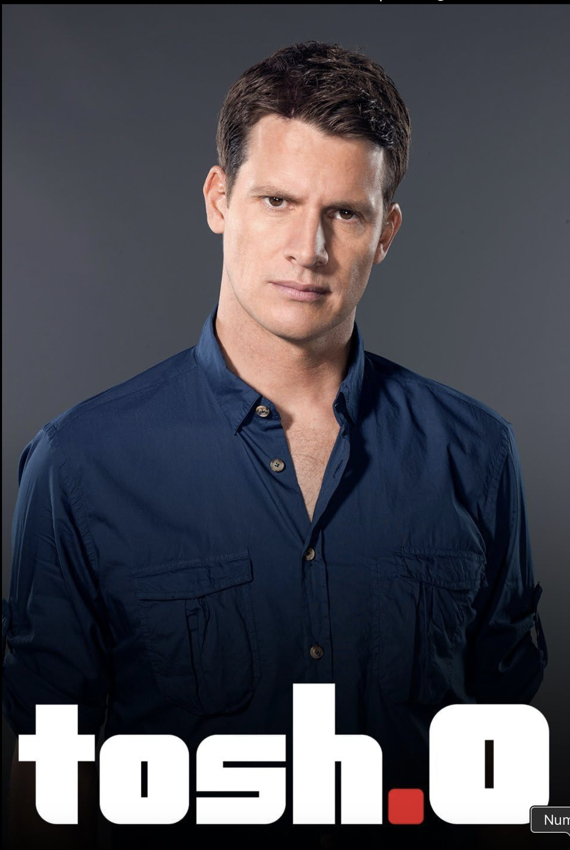 A man with dark hair wearing a navy blue button-up shirt, looking directly at the camera against a gray background. Part of the text 'tosh.o' is visible at the bottom of the image.
