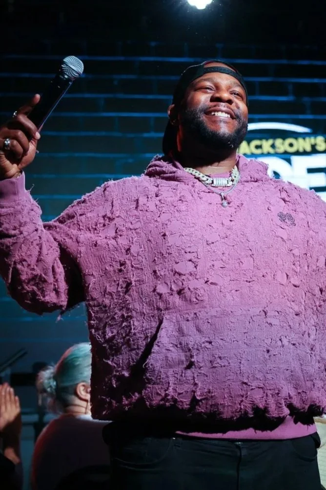 A man smiling on stage holding a microphone with a dark background and neon lights, wearing a purple textured hoodie and a baseball cap backward.