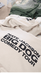 Clothes laid out on a bed, including a gray hoodie and a beige t-shirt with text about Nate Jackson's comedy tour.