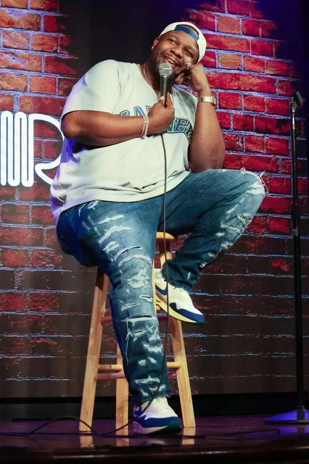 A man sitting on a wooden stool on stage, holding a microphone, with a red brick wall background and a partial neon sign.