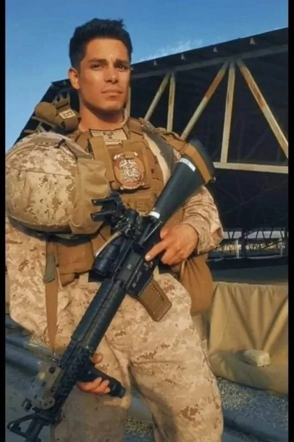 A soldier in desert camouflage military uniform holding a rifle with a scope, standing outdoors near a structure with a metal framework.