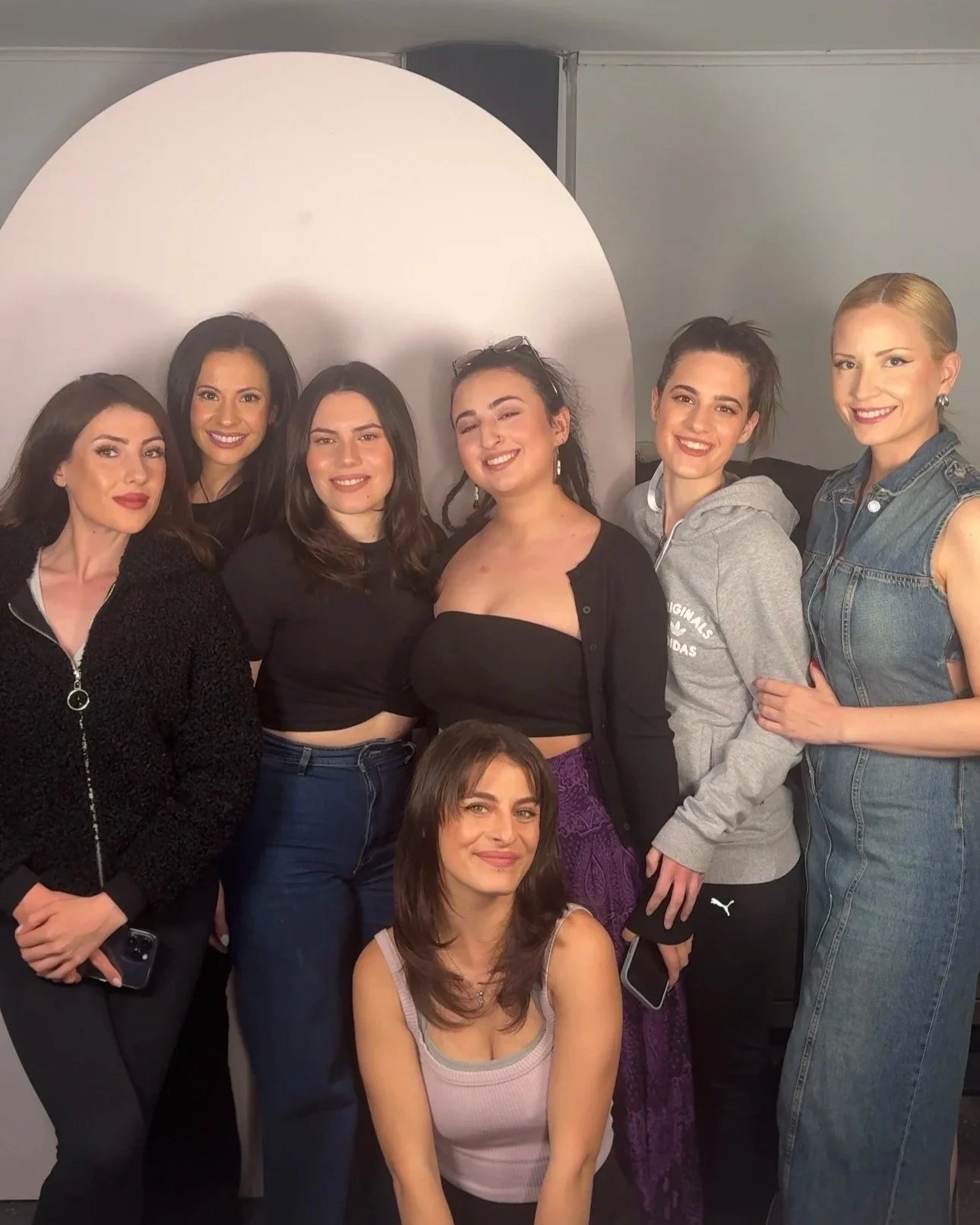 Group of seven women standing and sitting in front of a backdrop with a circular light. They are smiling and posing for a photo.