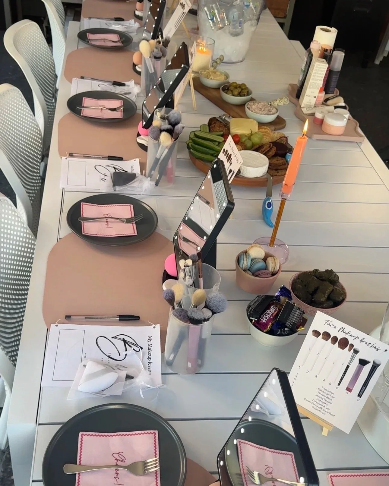 A table set for a catering event with pink placemats, black plates, and utensils wrapped in pink napkins. The table includes makeup brushes, beauty products, and snacks such as cookies, macarons, grapes, pickles, and chocolate bars. There are also candles and mirrors on the table.