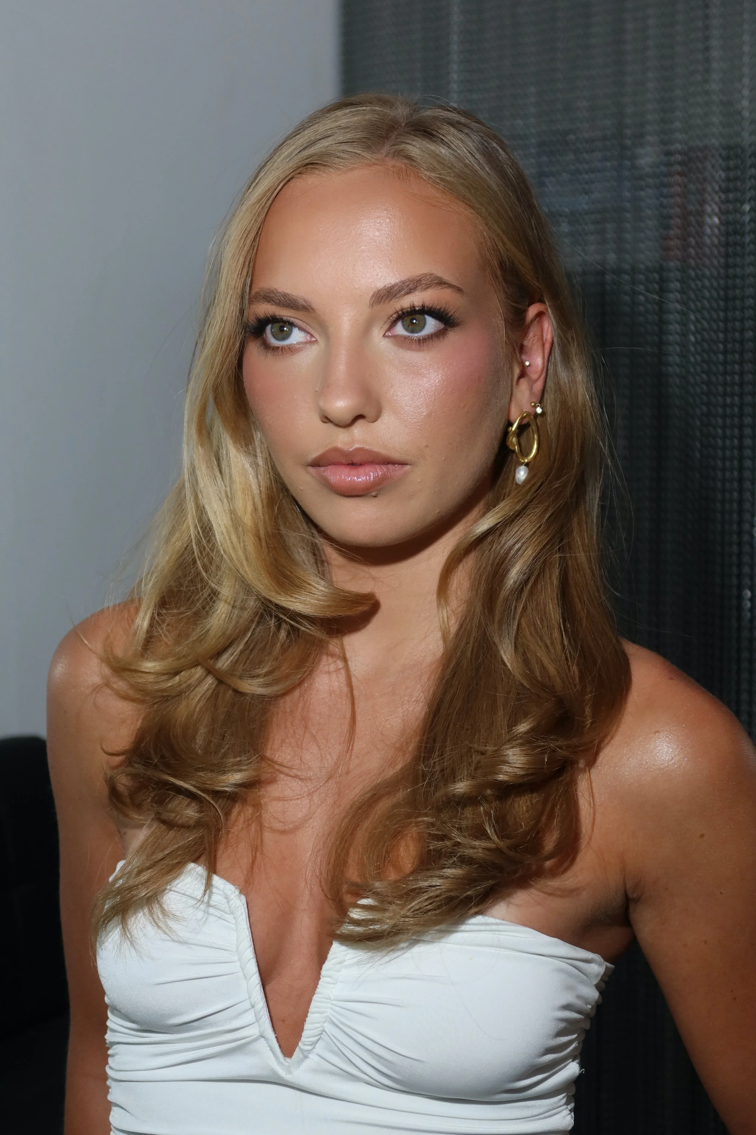 Close-up of a young woman with long, wavy blonde hair wearing a white dress with a plunging neckline, gold earrings with pearl accents, looking slightly to the side.