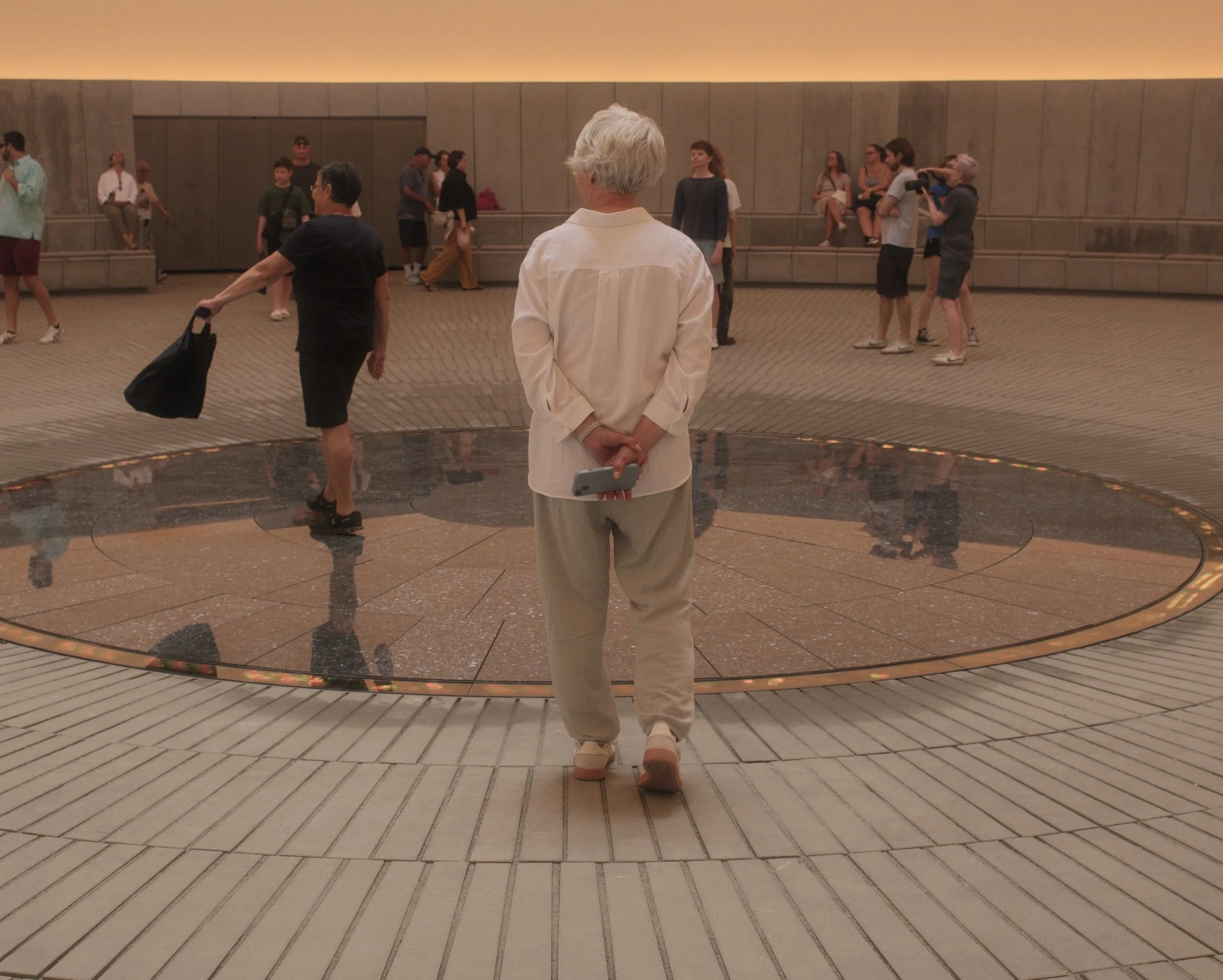 A woman with gray hair dressed in beige standing with her back to the camera inside a spacious, well-lit terminal or lobby area. The area has a curved, decorative floor and a large wall behind her with several people sitting and standing, some with c