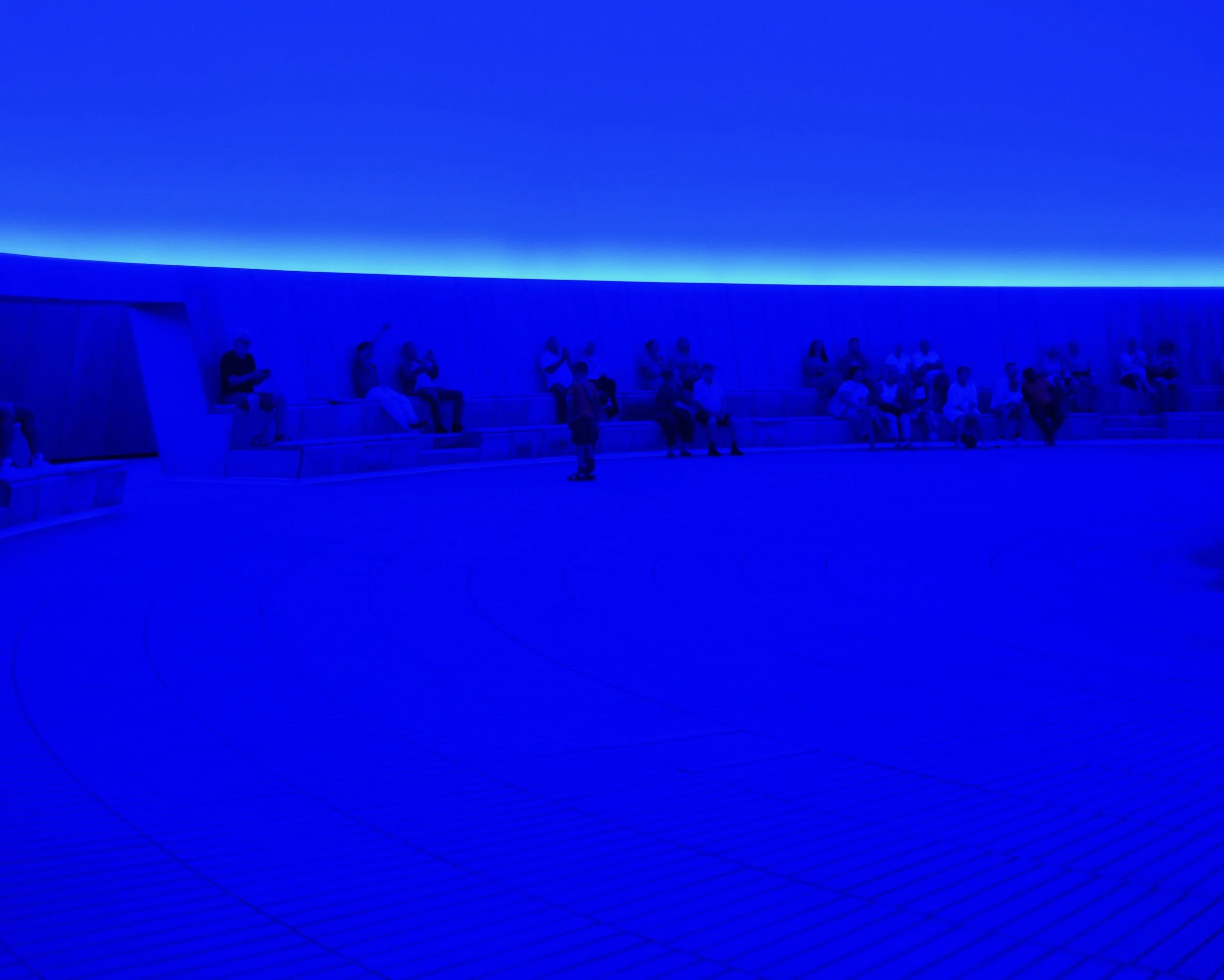Room with blue lighting, rows of seating, and people sitting and standing, some taking photos or looking around.