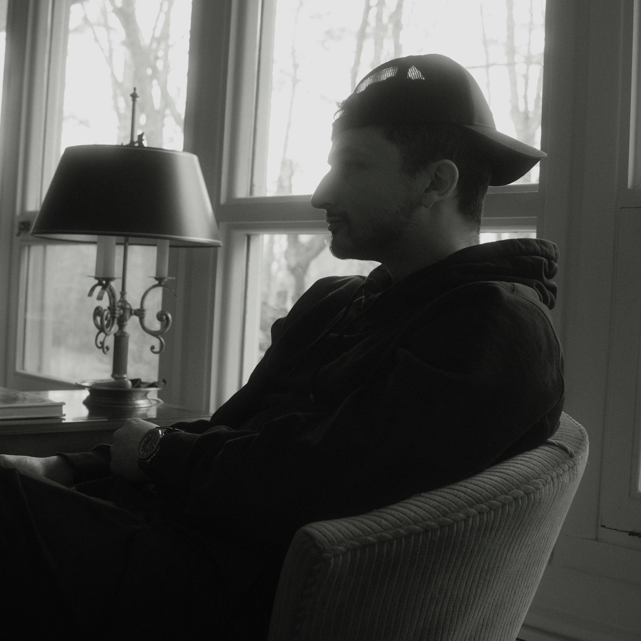 Side view of a person wearing a baseball cap sitting in a room with large windows, a lamp, and a table with books.