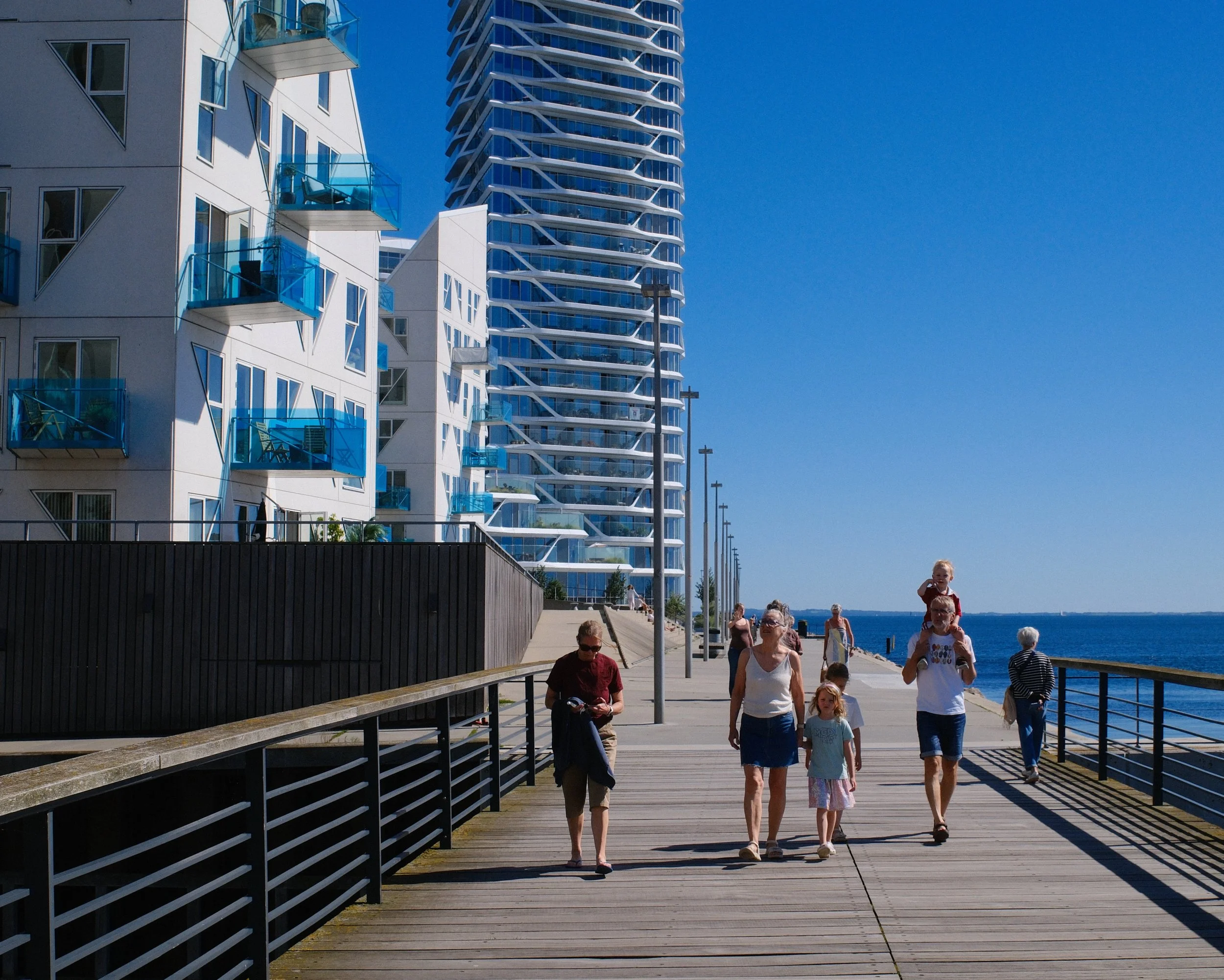 People walking along a wooden boardwalk by the water with modern high-rise buildings in the background on a sunny day.