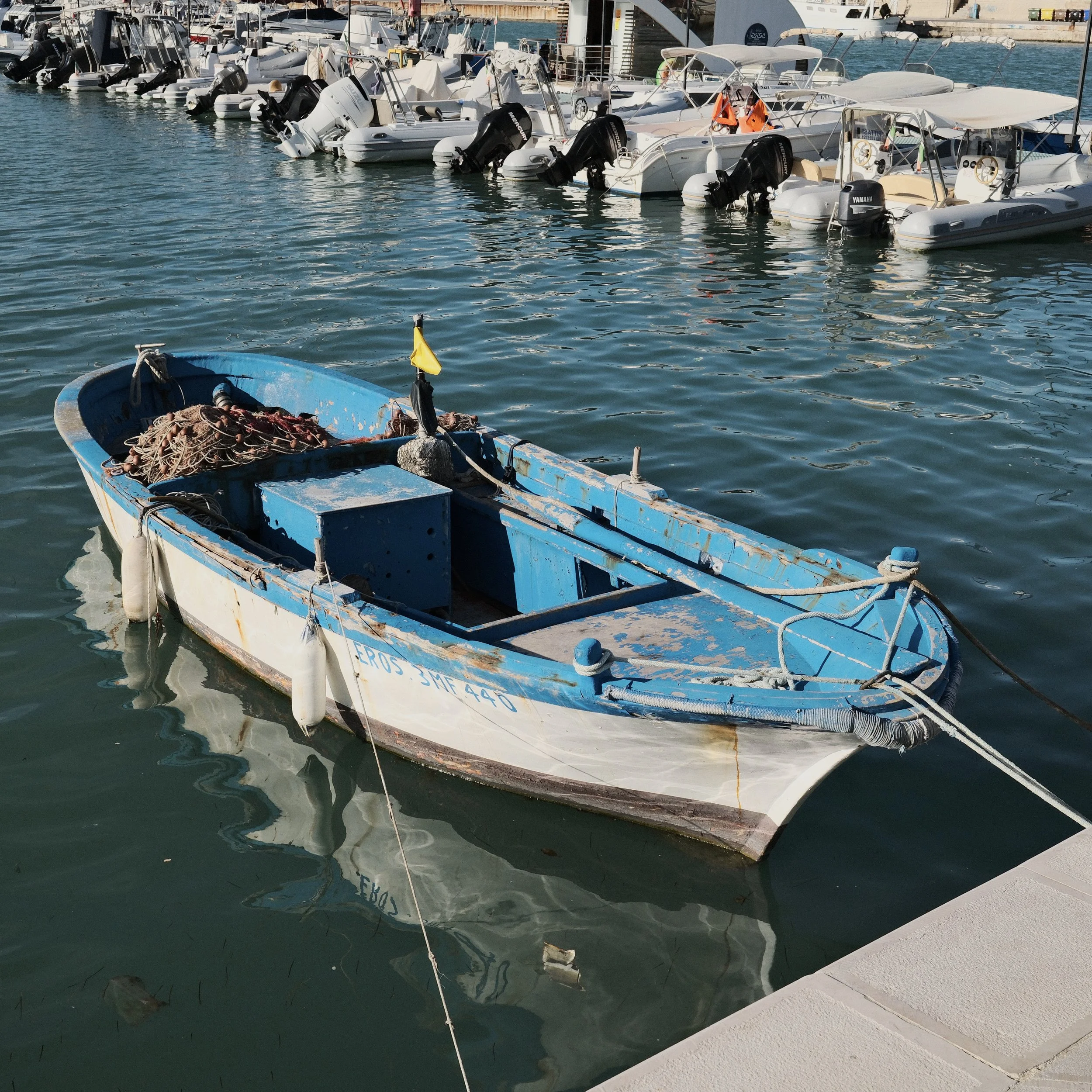 A small blue boat filled with tangled ropes floating in the water near a dock, with a row of white boats with outboard motors moored in the background.