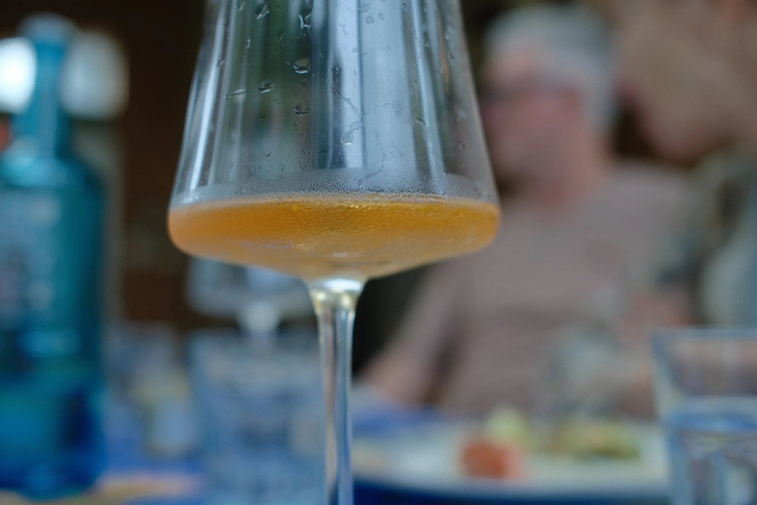 A wine glass with orange-colored beverage, labels on bottles in the background, and condensation on the glass.
