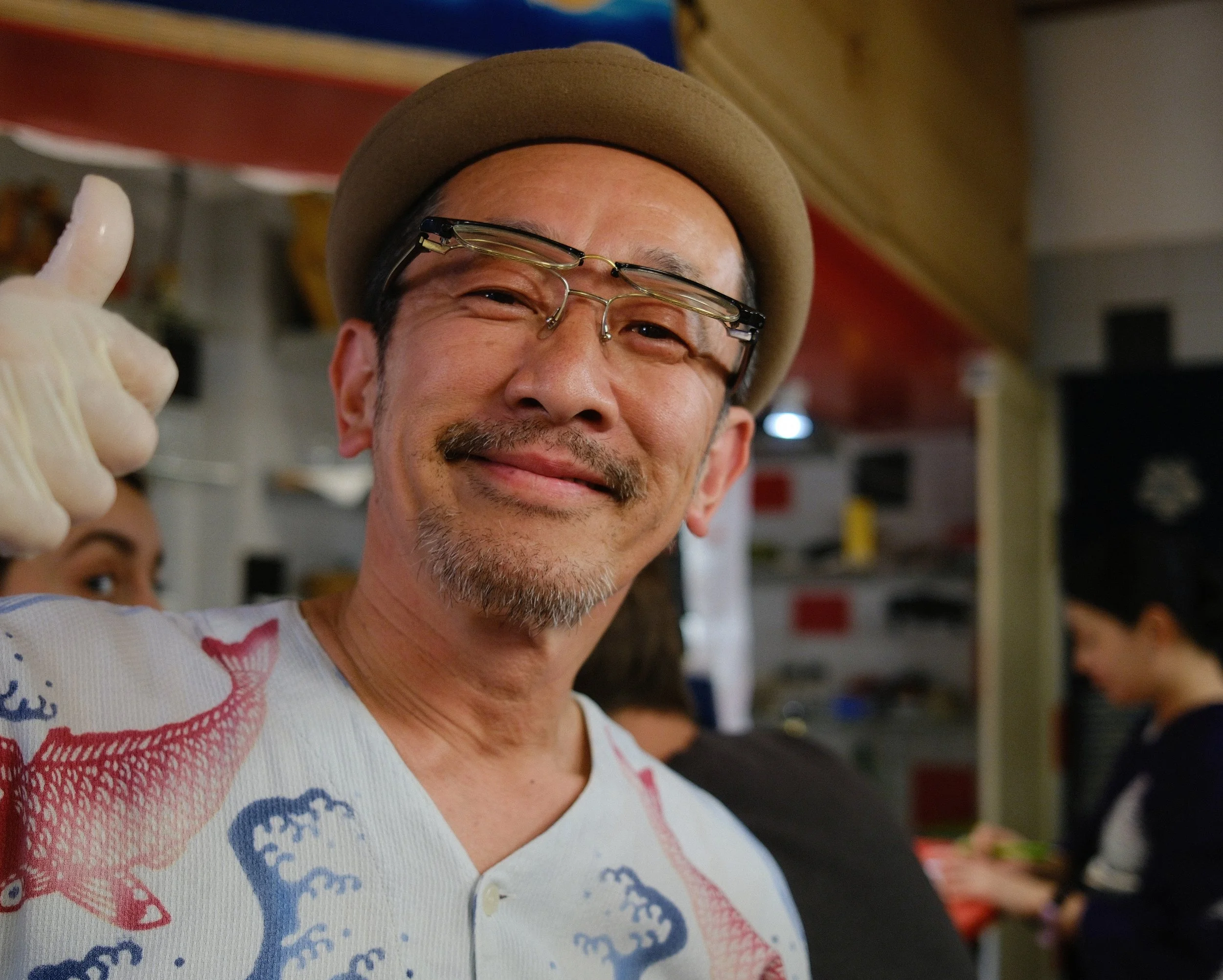 A smiling man with glasses and a tan hat giving a thumbs-up. He is wearing a colorful shirt with red, blue, and white patterns. The background shows a store interior with shelves and other people.