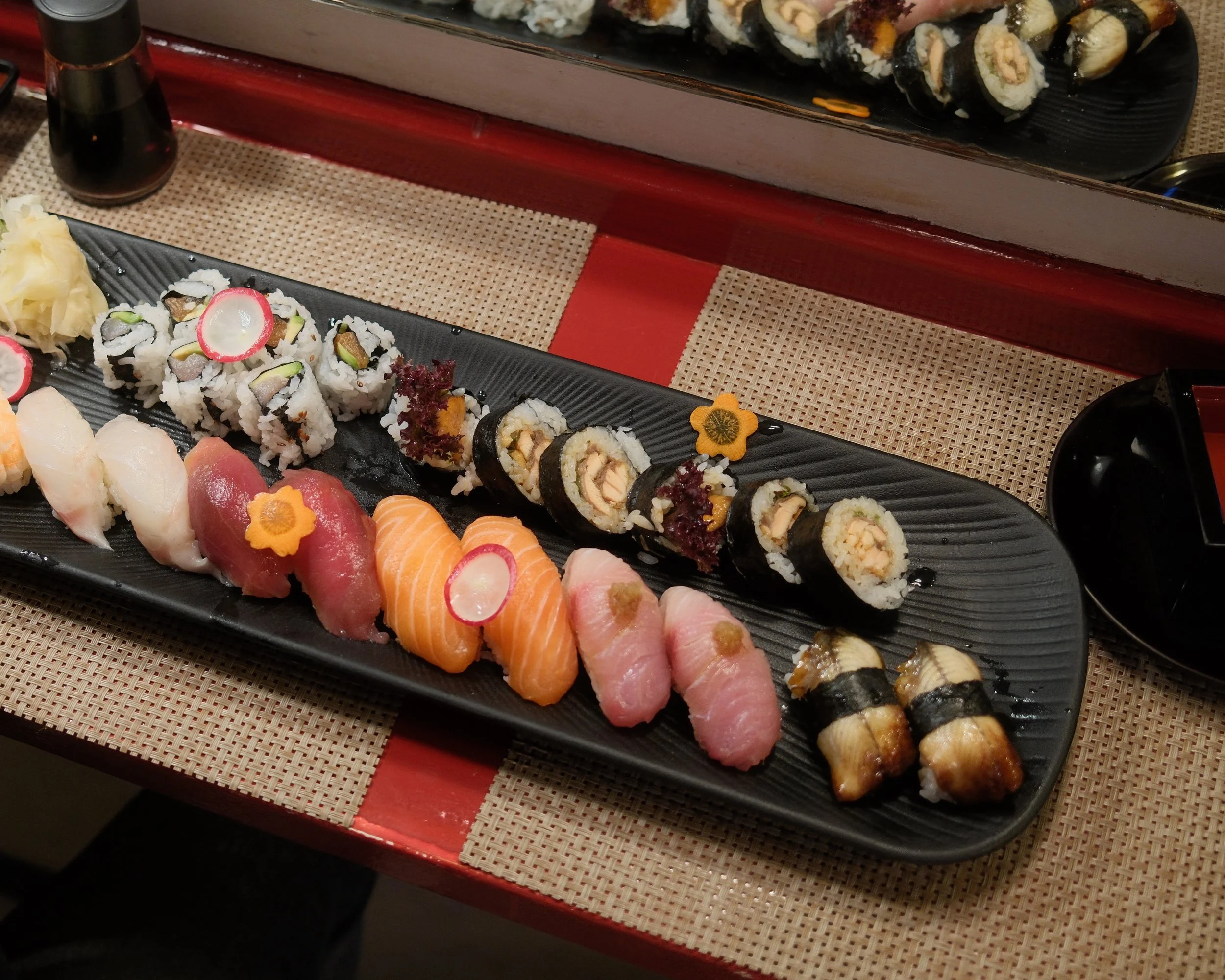 A black sushi platter with assorted sushi rolls, sashimi, and garnishes, placed on a woven placemat on a table.
