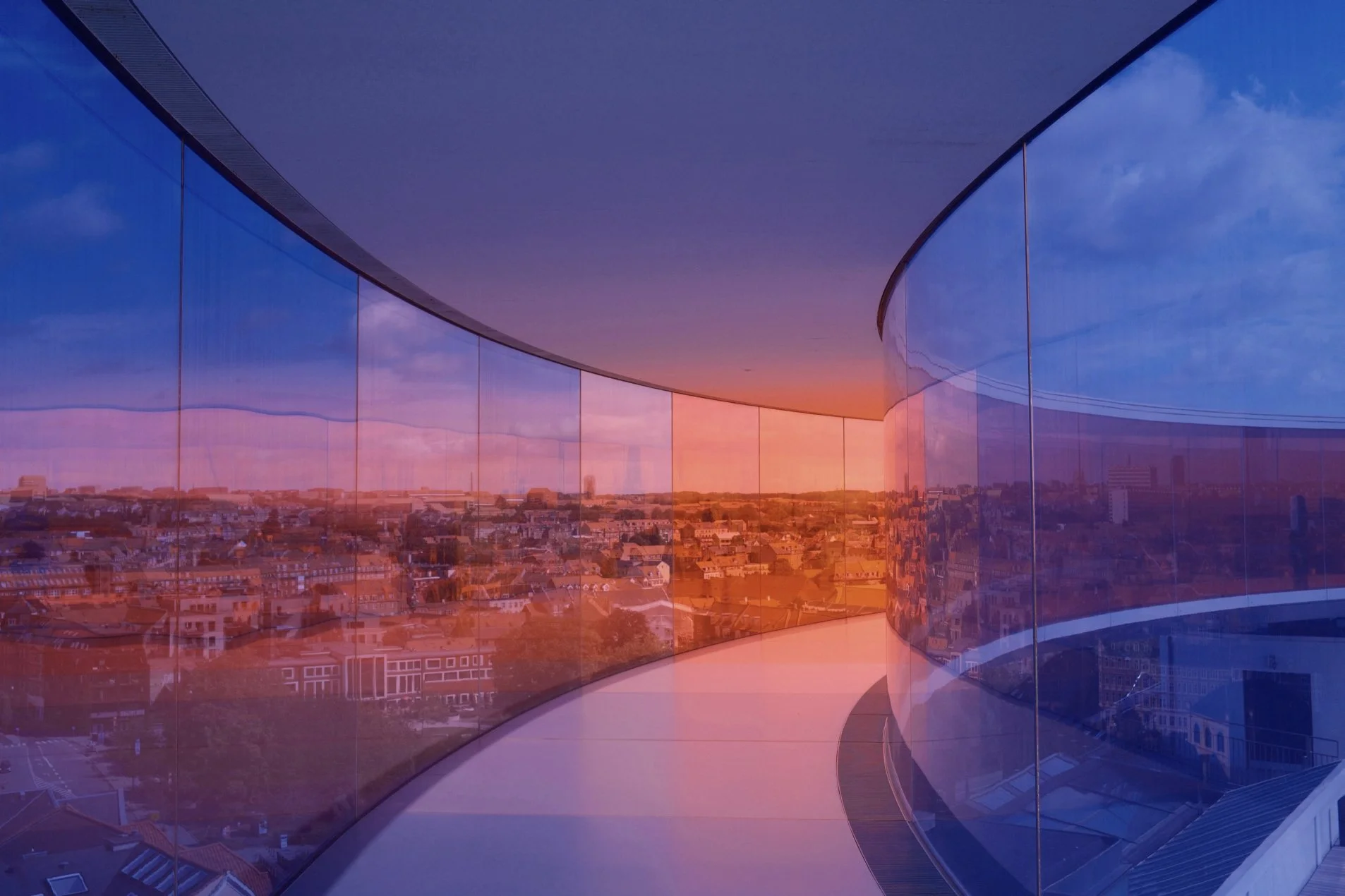 A panoramic view of a city through curved, reflective glass windows during sunset, with colors transitioning from deep blue to orange and pink in the sky.