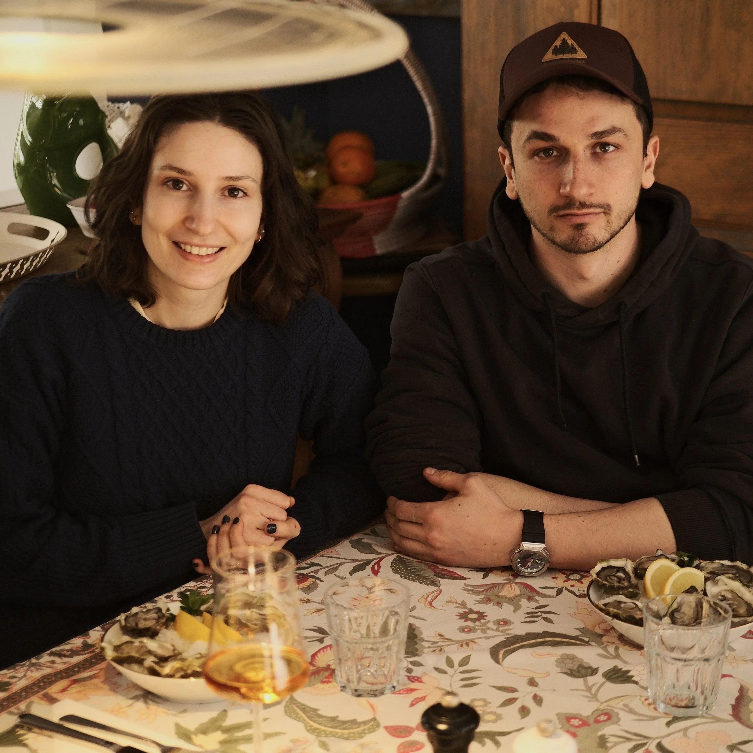Two people sitting at a table with seafood dishes and drinks. A woman on the left is smiling and a man on the right is looking at the camera. The table has a patterned tablecloth, and there are plates of oysters, lemon wedges, and glasses of wine.