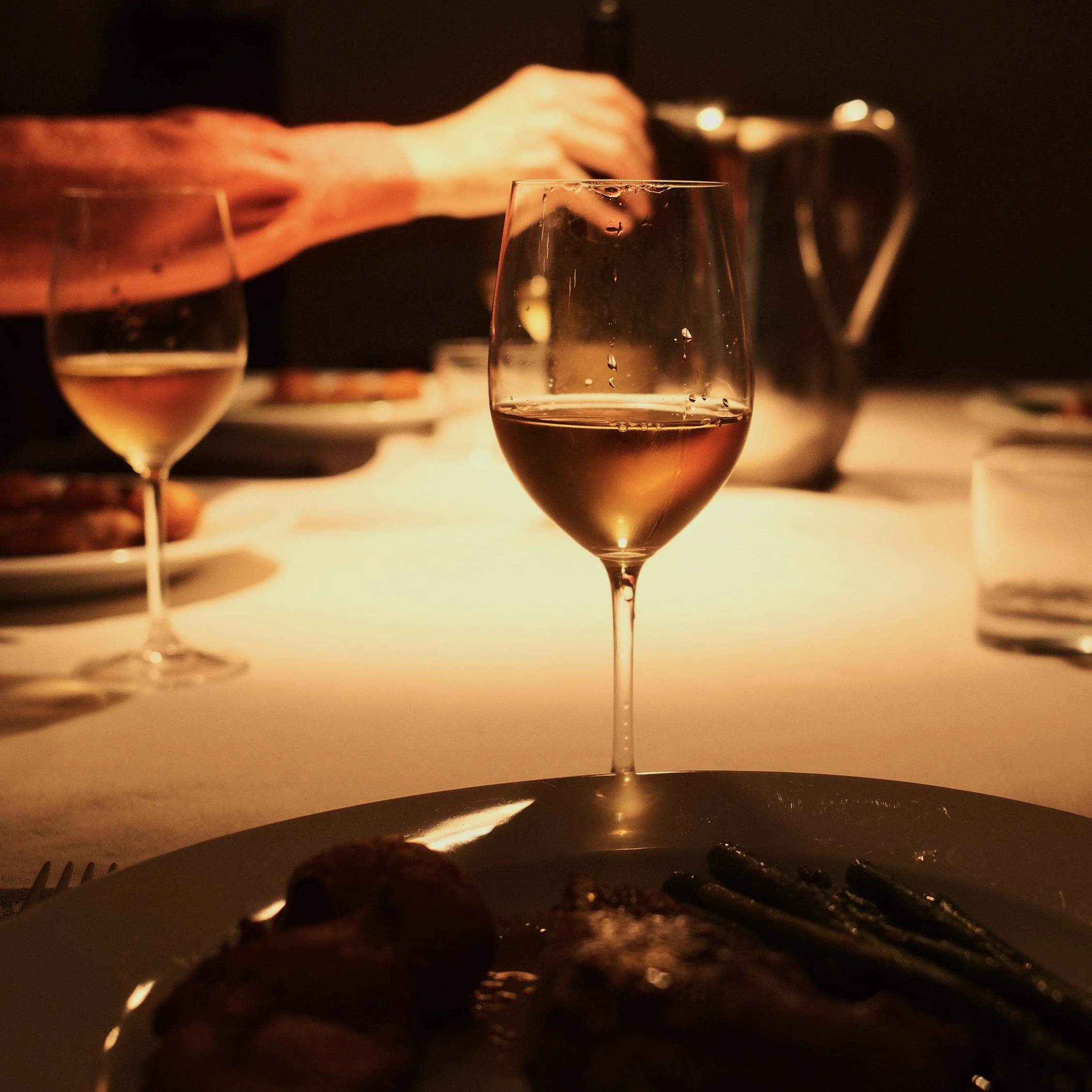 Close-up of a glass of white wine on a table with a blurred background of a person pouring a drink, warm lighting creating a cozy atmosphere.