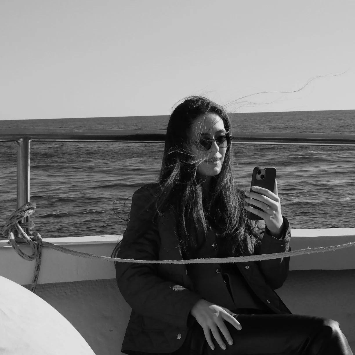 A woman consulting her mobile phone, sitting on the deck of a boat with the sea in the background.