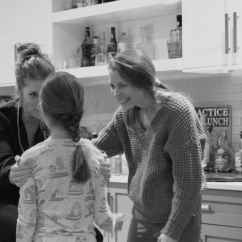 Two women smiling and talking to a young girl in a kitchen with shelves of bottles and cabinets in the background.