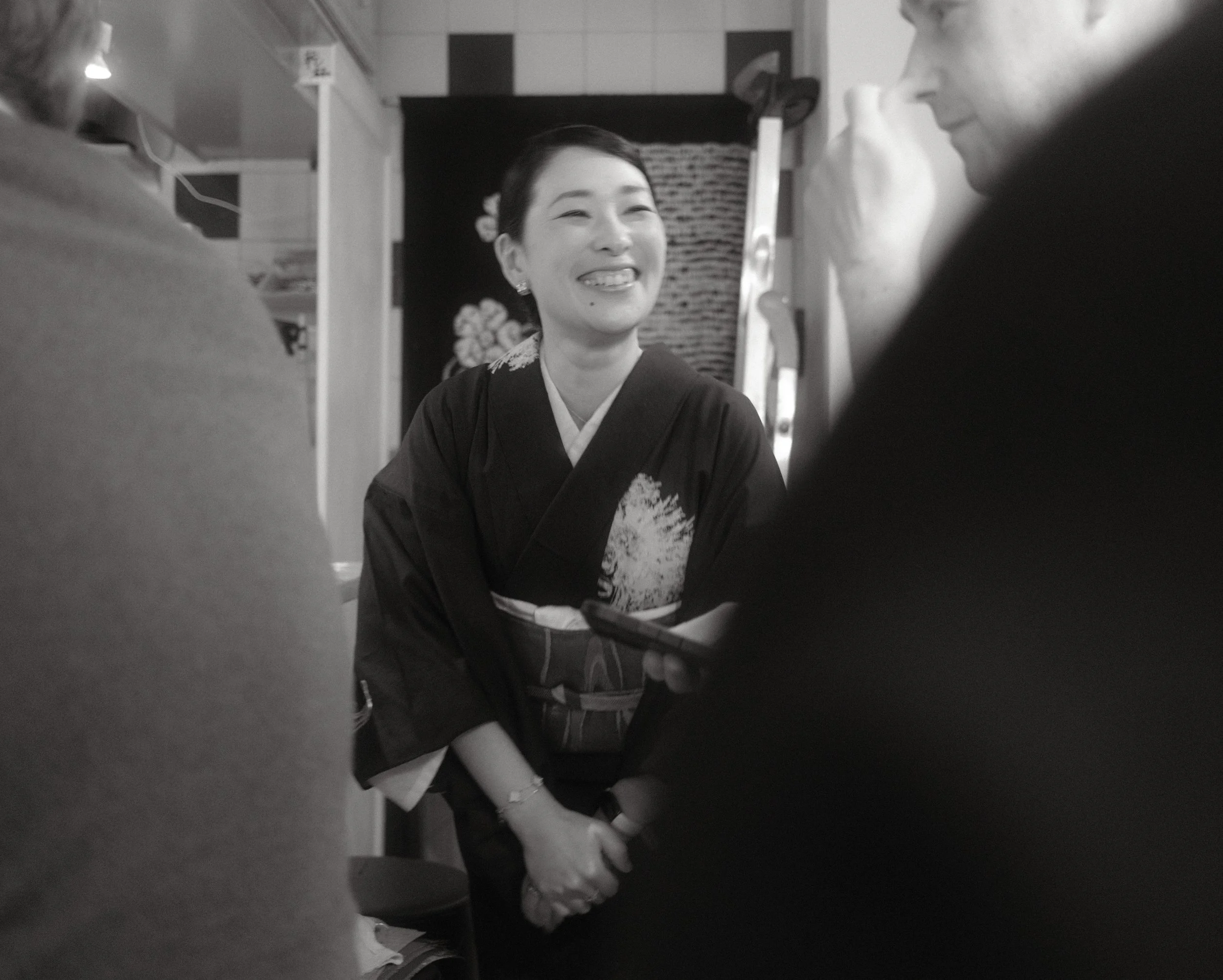 Black and white photo of a young smiling woman dressed in traditional Japanese kimono, sitting with her eyes closed, while a man in a suit looks at her, both framed by a blurry foreground. Indoor setting with decorative wall elements in the backgroun