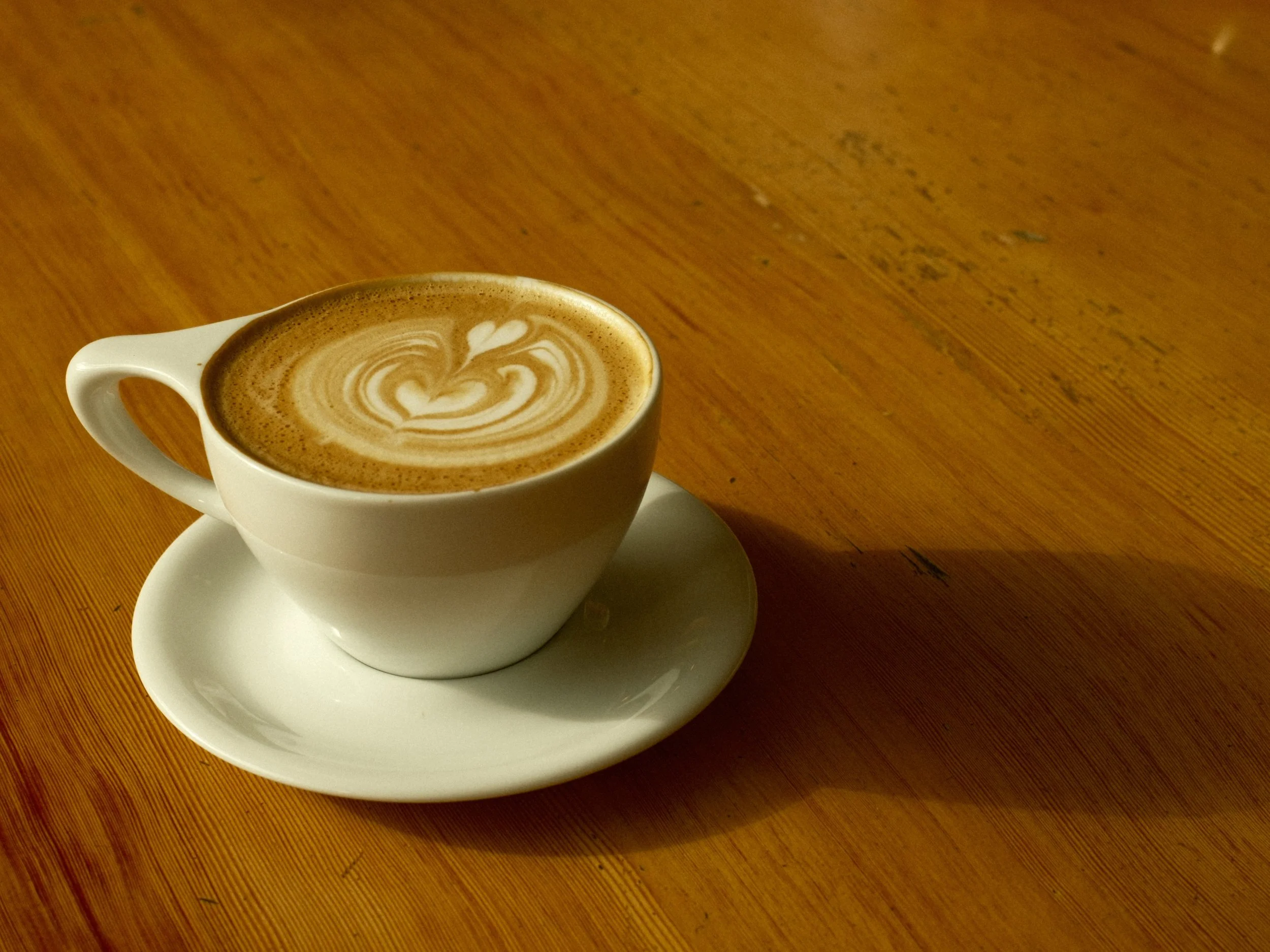 A white ceramic cup of coffee with latte art featuring a heart and leaf design, placed on a matching saucer on a wooden table.