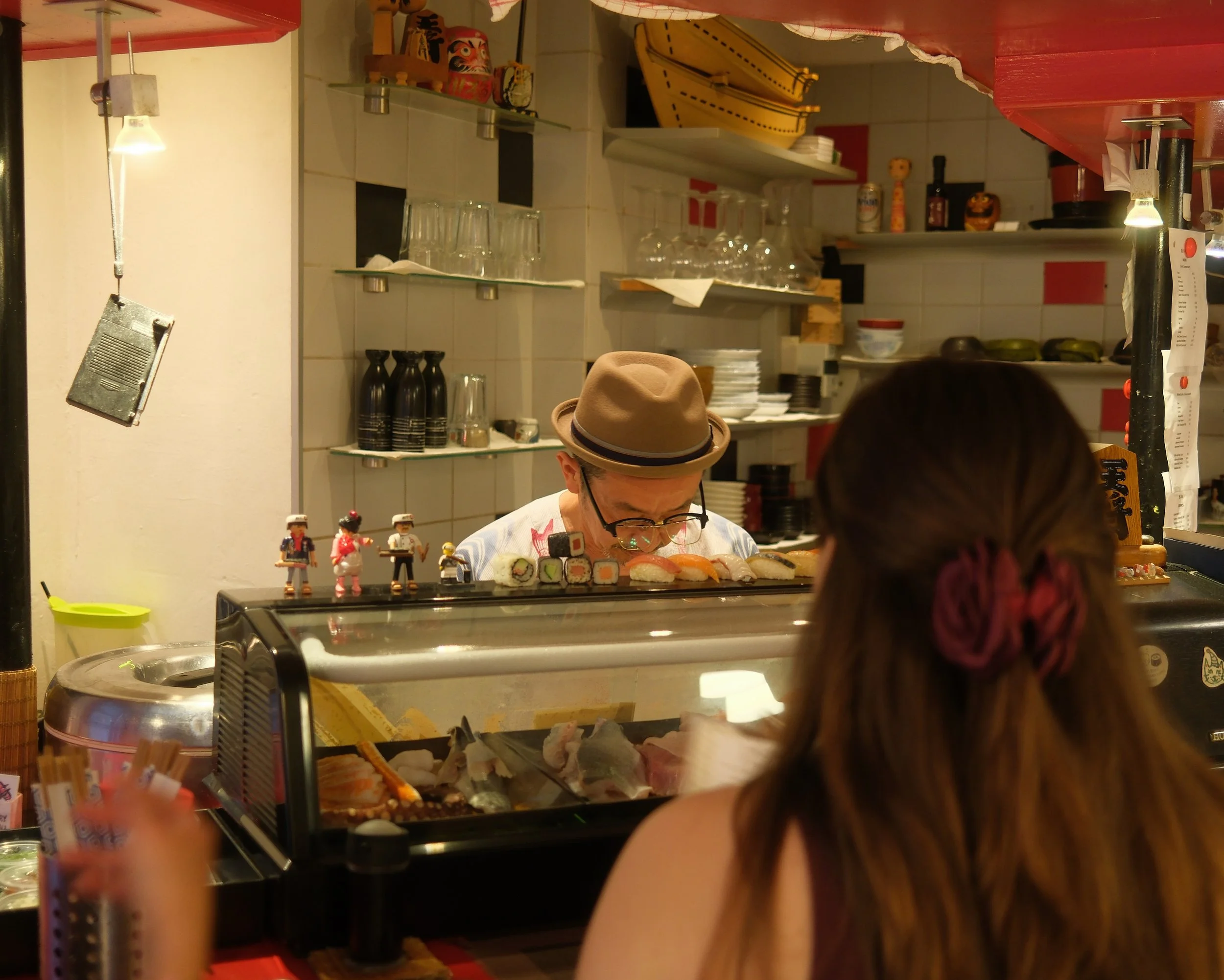 People ordering sushi at a counter in a restaurant, with various sushi rolls visible on the counter and Japanese decorative items on the shelves in the background.