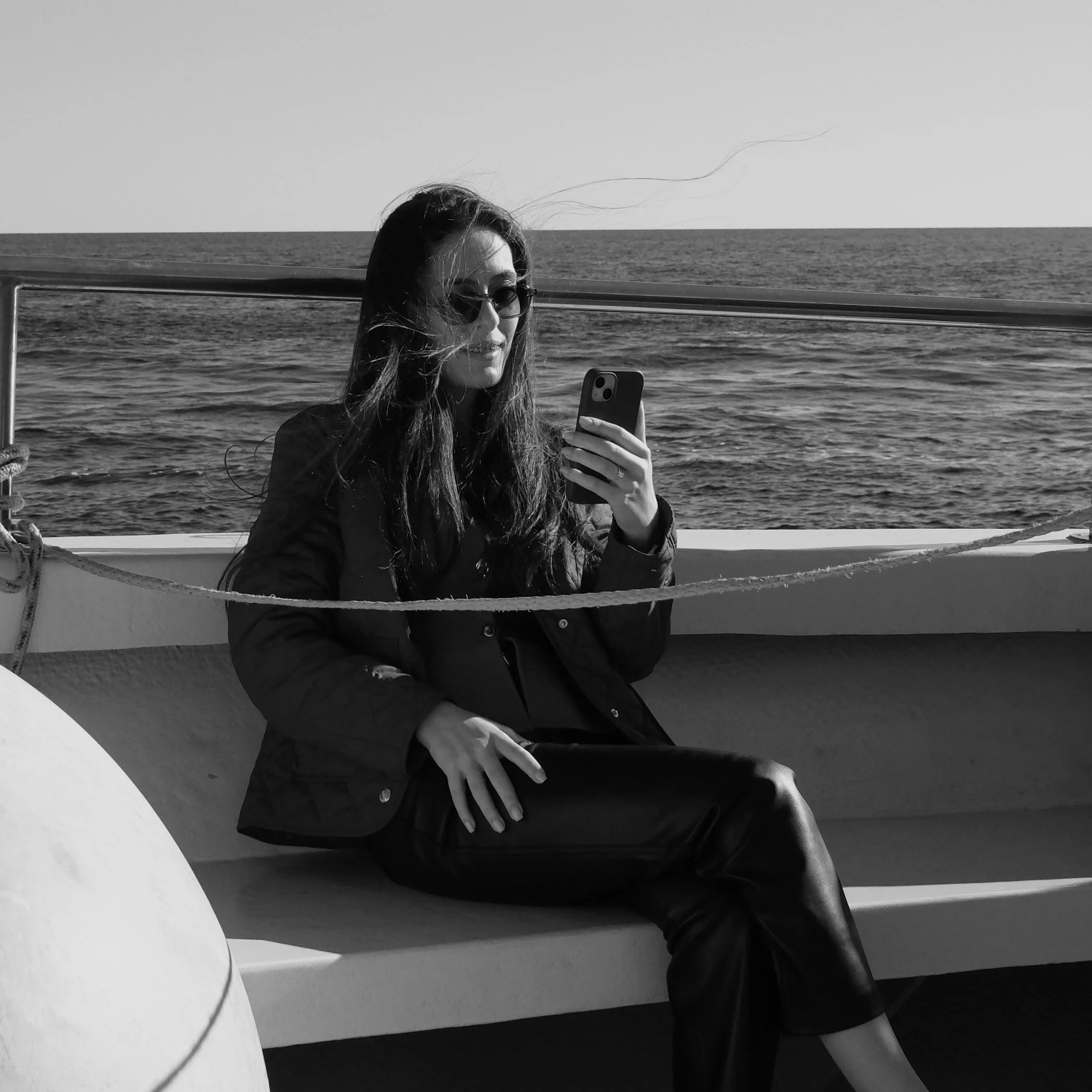 A woman with long hair and sunglasses takes a selfie on a boat, with an ocean and clear sky in the background.